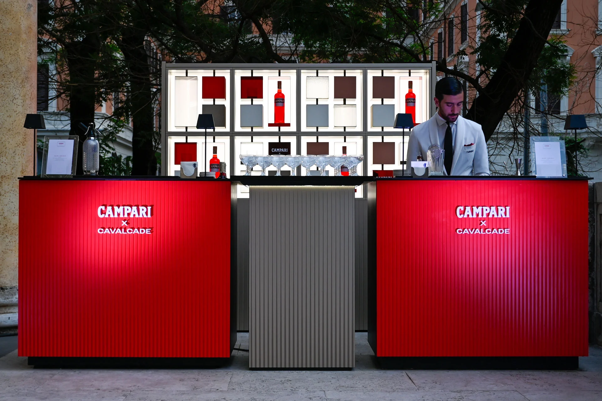 Two red Campari x Cavalcade bar counters in front of a backlit shelf, with a bartender – Campari Cavalcade, Rome, Italy, 2023 – event design