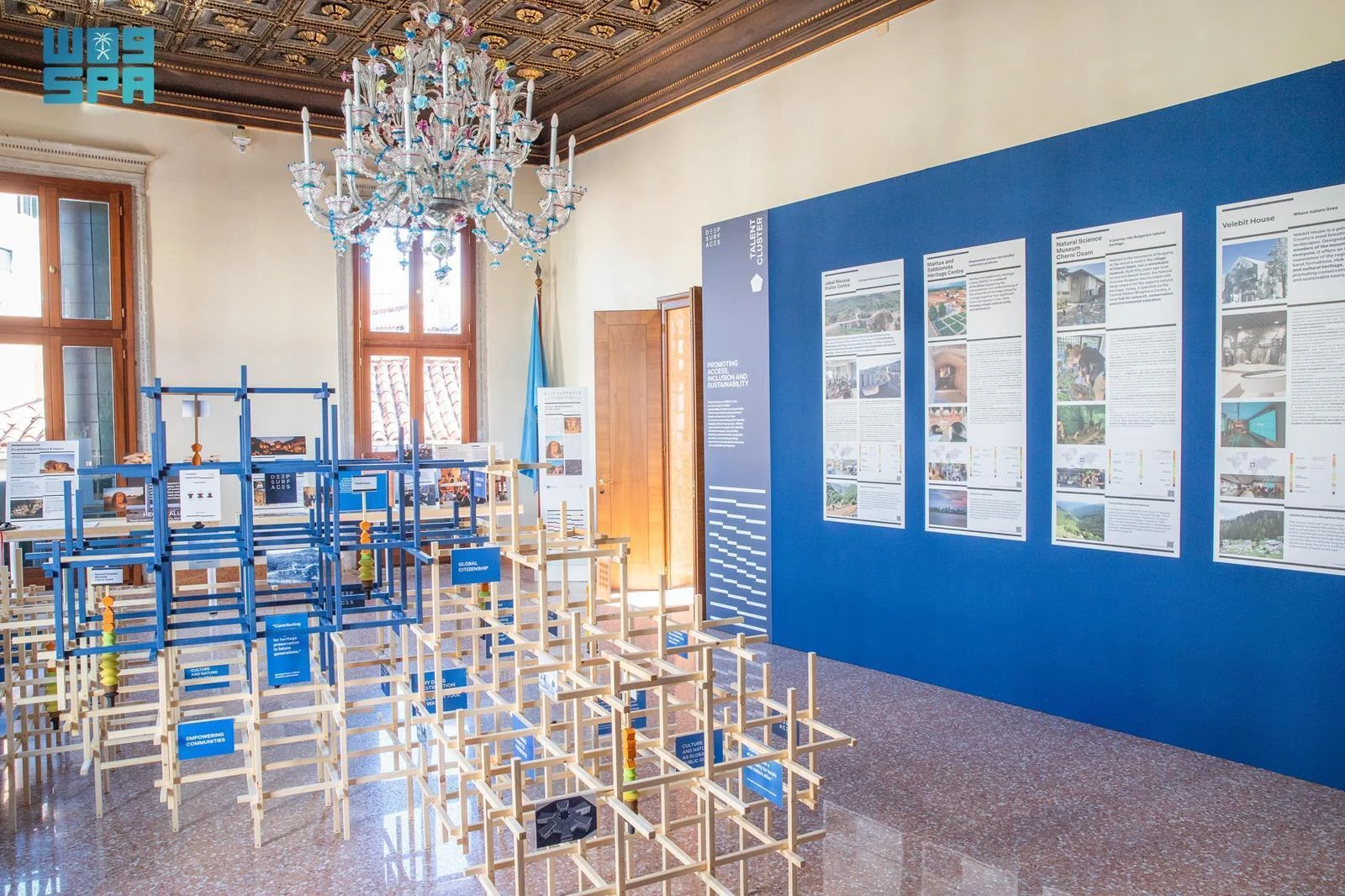 Exhibition room with blue and natural wooden lattice structures under a Murano chandelier, project panels on a blue wall – Deep Surfaces at Palazzo Zorzi, Venice, Italy, 2025 – exhibition design