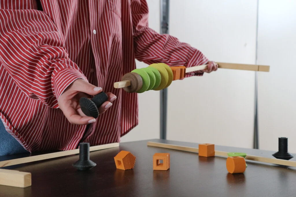 Hands threading green and brown 3D-printed pieces onto a wooden stick, other pieces on the table – Deep Surfaces at Palazzo Zorzi, Venice, Italy, 2025 – exhibition design
