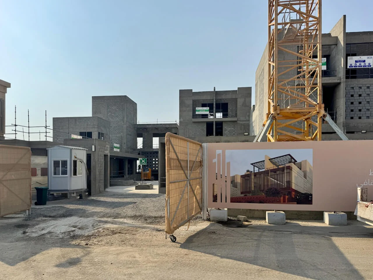 Construction site entrance with concrete villa shells, a tower crane and a hoarding showing the project render – Radic Residences, Jeddah, Saudi Arabia, 2025 – architecture, under construction