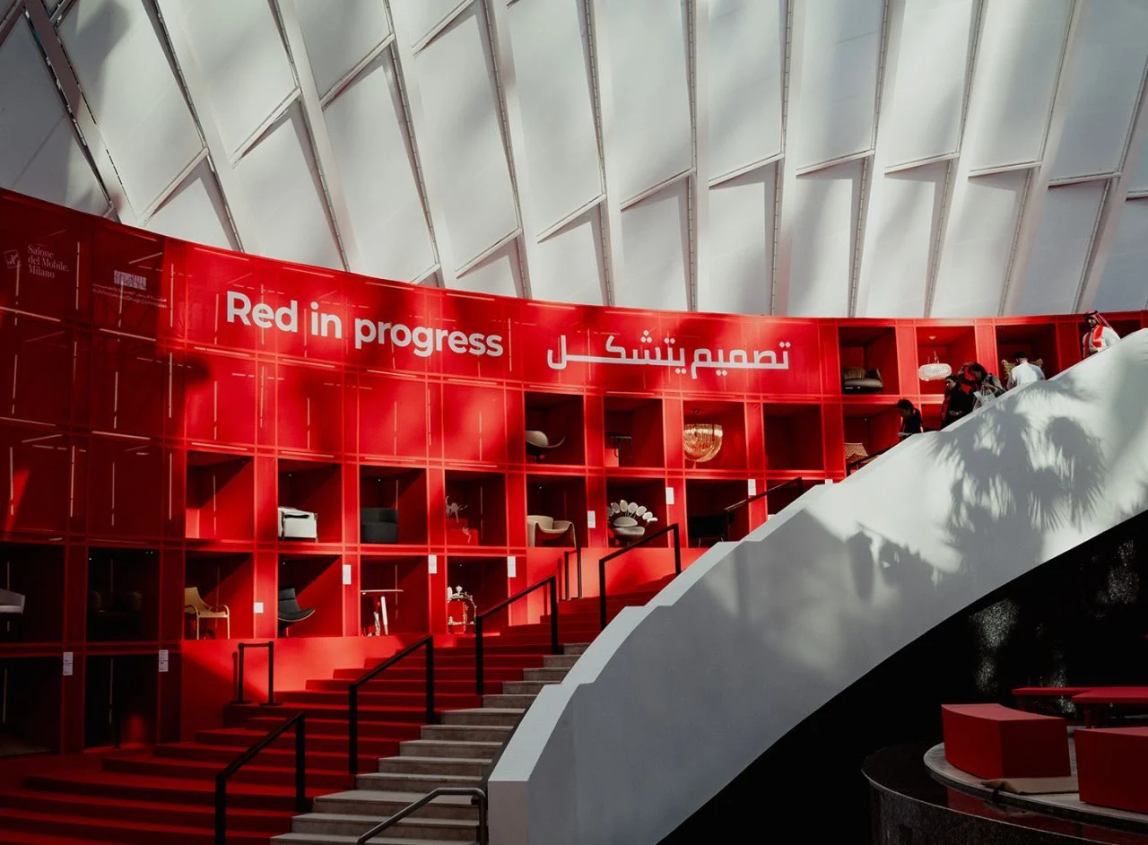 Red shelving wall reading Red in progress, displaying furniture beside a red staircase under a white canopy – Red in Progress: Salone del Mobile Meets Riyadh, Saudi Arabia, 2025 – exhibition design