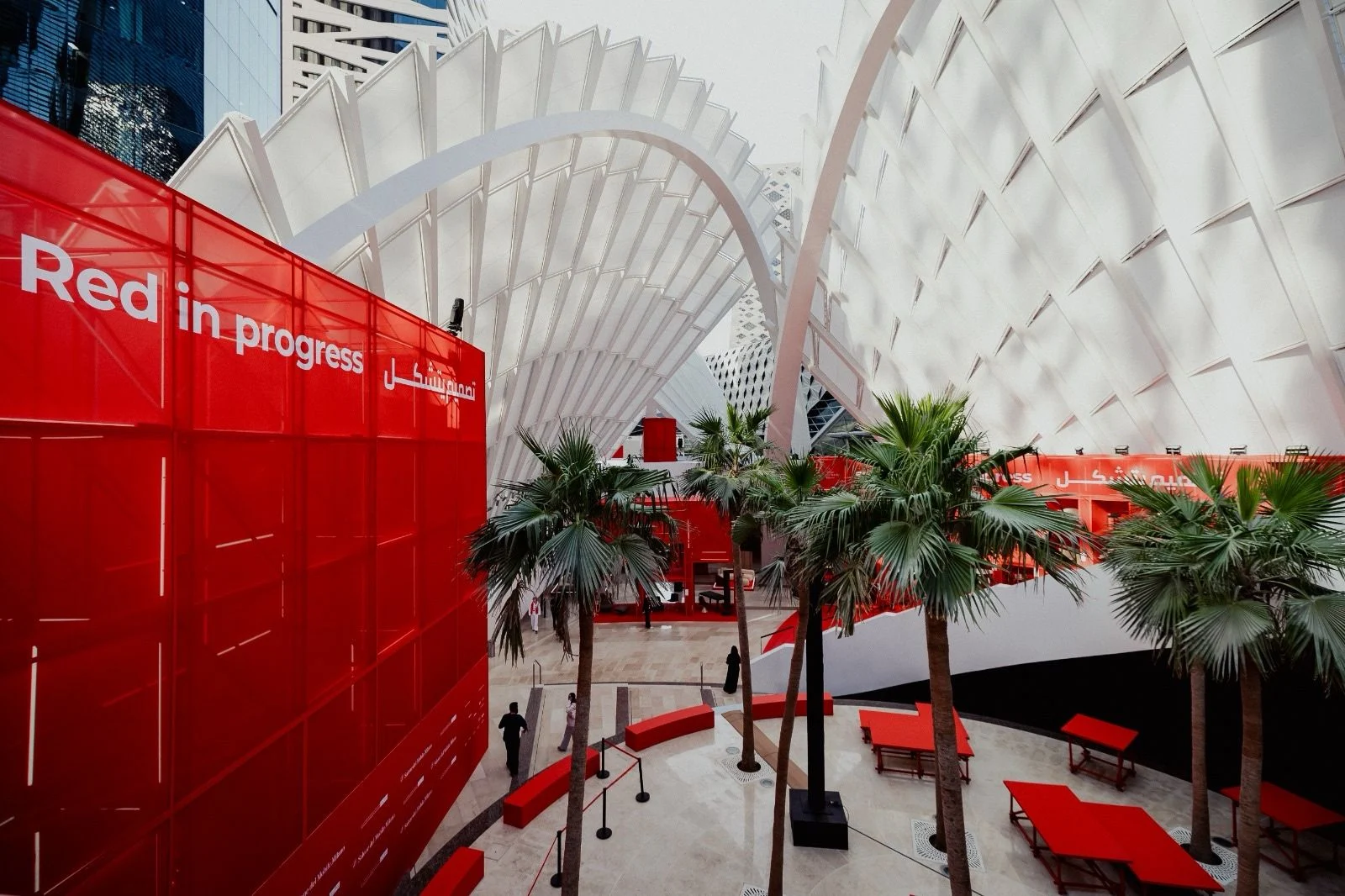 Red in progress installation: a red glass volume and red tables among palms under white ribbed canopies – Red in Progress: Salone del Mobile Meets Riyadh, Saudi Arabia, 2025 – exhibition design