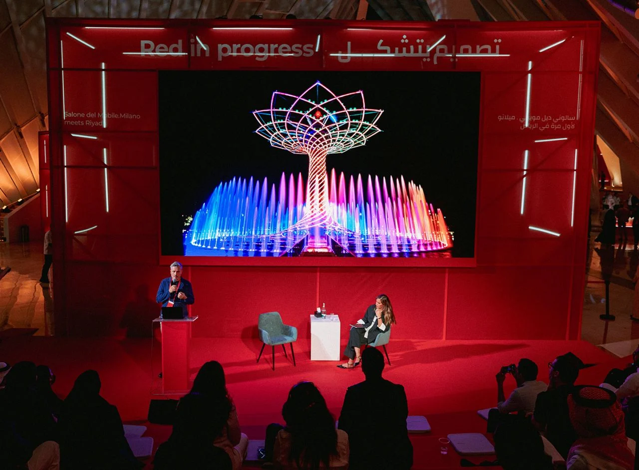 Talk on the red stage, a screen behind showing a lit tree sculpture over fountains, audience in front – Red in Progress: Salone del Mobile Meets Riyadh, Saudi Arabia, 2025 – exhibition design