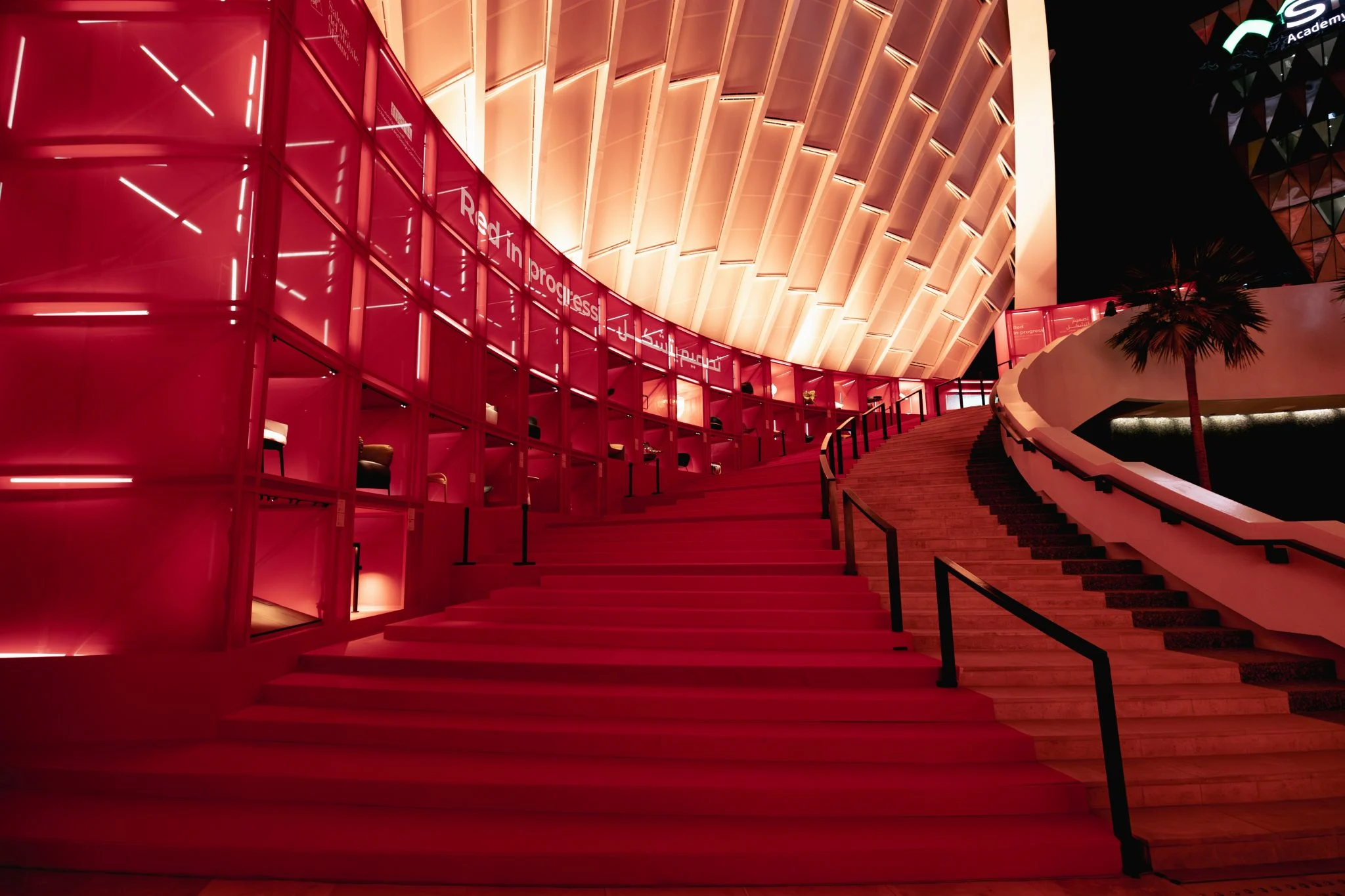 Night view of the red staircase rising along illuminated display shelves under the curved canopy – Red in Progress: Salone del Mobile Meets Riyadh, Saudi Arabia, 2025 – exhibition design