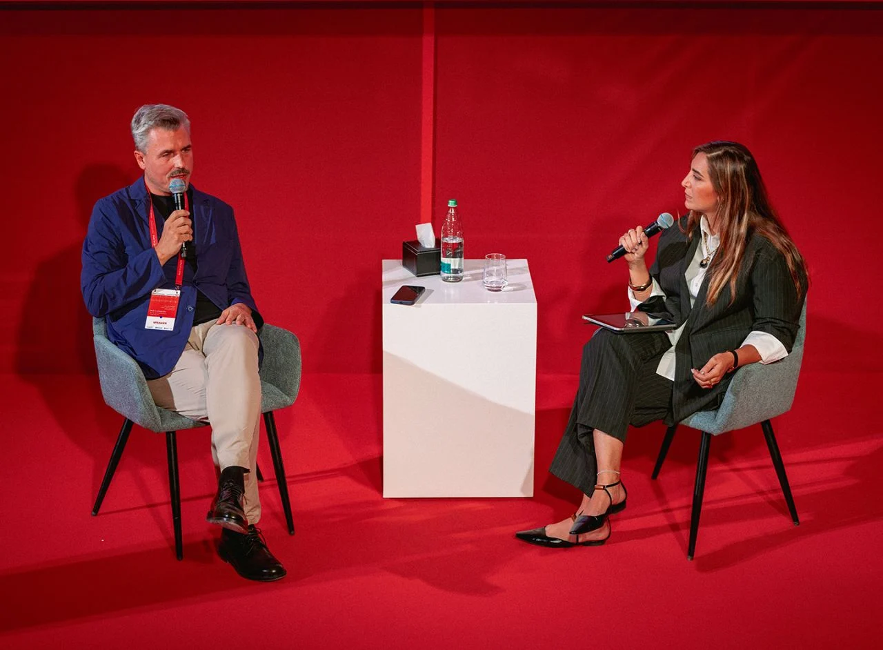 Two speakers seated in conversation with microphones on a red stage, a white plinth between them – Red in Progress: Salone del Mobile Meets Riyadh, Saudi Arabia, 2025 – exhibition design