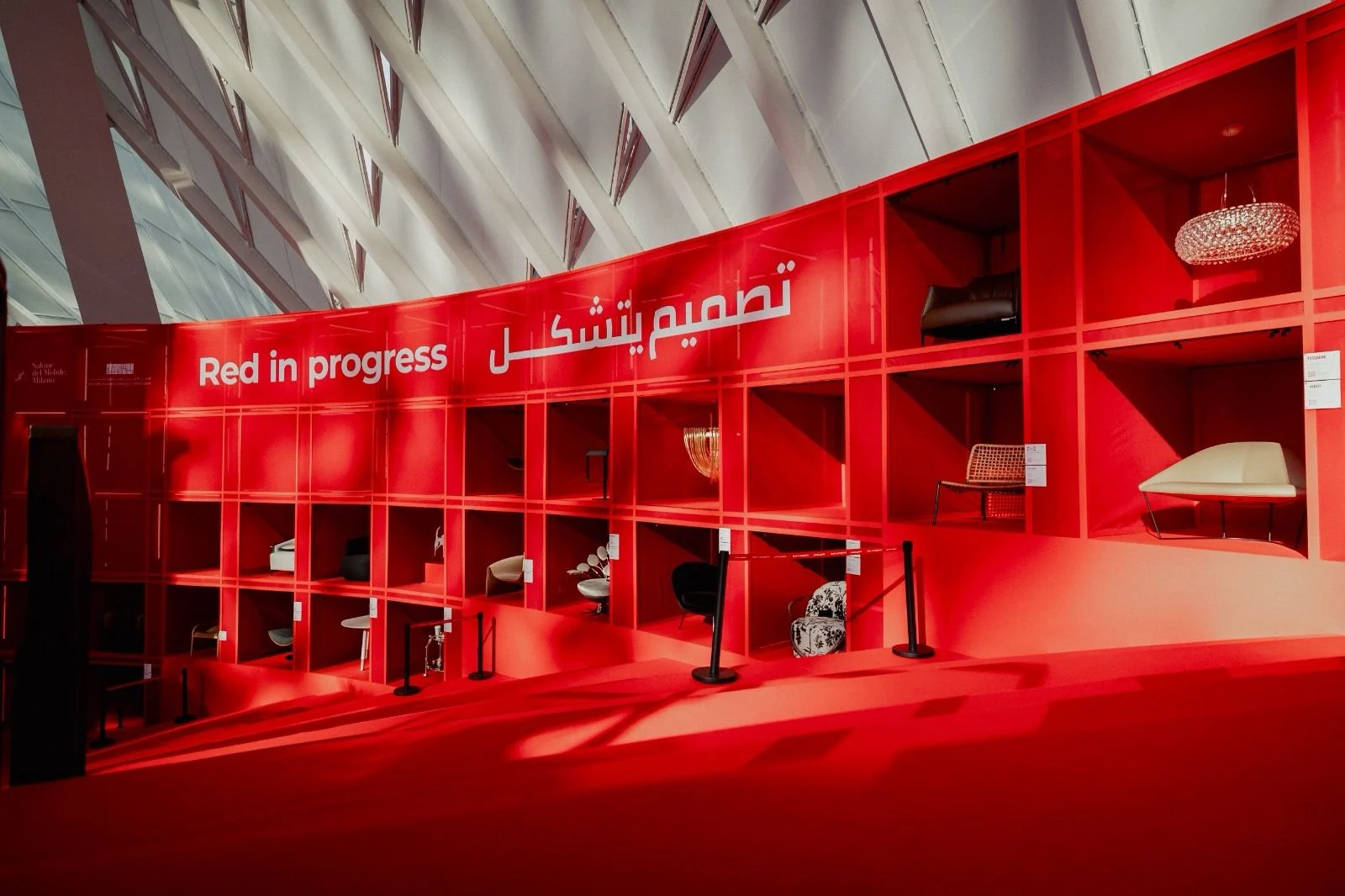 Curved red display wall lettered Red in progress in English and Arabic, chairs and lamps in its niches – Red in Progress: Salone del Mobile Meets Riyadh, Saudi Arabia, 2025 – exhibition design