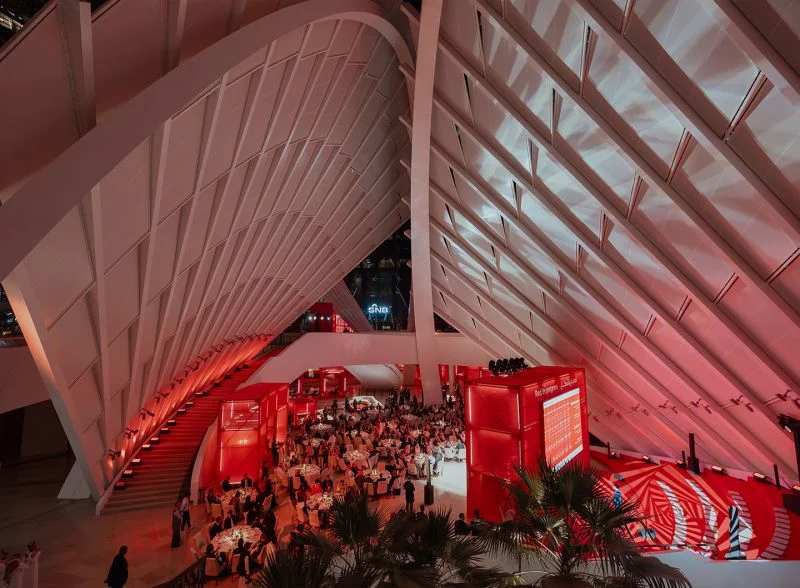 Gala dinner tables beneath the red-lit ribbed canopy, seen from above beside a red staircase – Red in Progress: Salone del Mobile Meets Riyadh, Saudi Arabia, 2025 – exhibition design