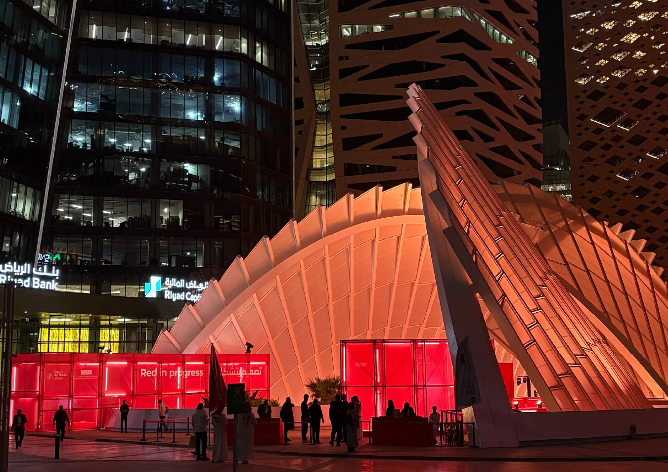 Night view of the glowing white canopy and red installation among office towers, visitors on the plaza – Red in Progress: Salone del Mobile Meets Riyadh, Saudi Arabia, 2025 – exhibition design