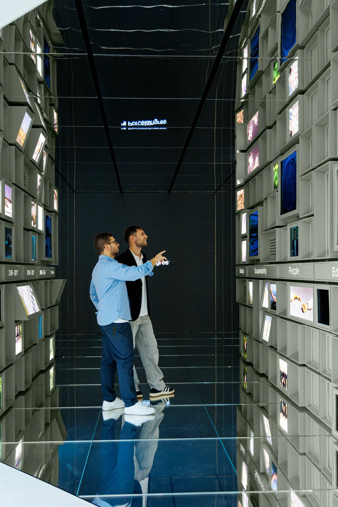 Two visitors inside the mirrored corridor between walls of cubes, one pointing at a screen – Porcelaingres Brand Narrative Space, Castellarano, Italy, 2025 – showroom design