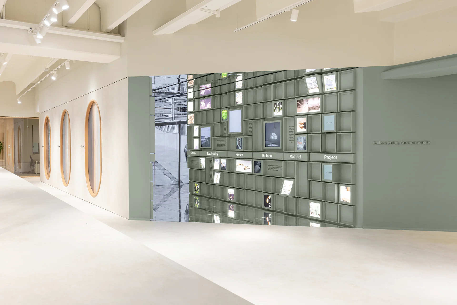 Showroom entrance to the mirrored brand space: a sage-green modular wall of screens and images beside oval niches – Porcelaingres Brand Narrative Space, Castellarano, Italy, 2025 – showroom design