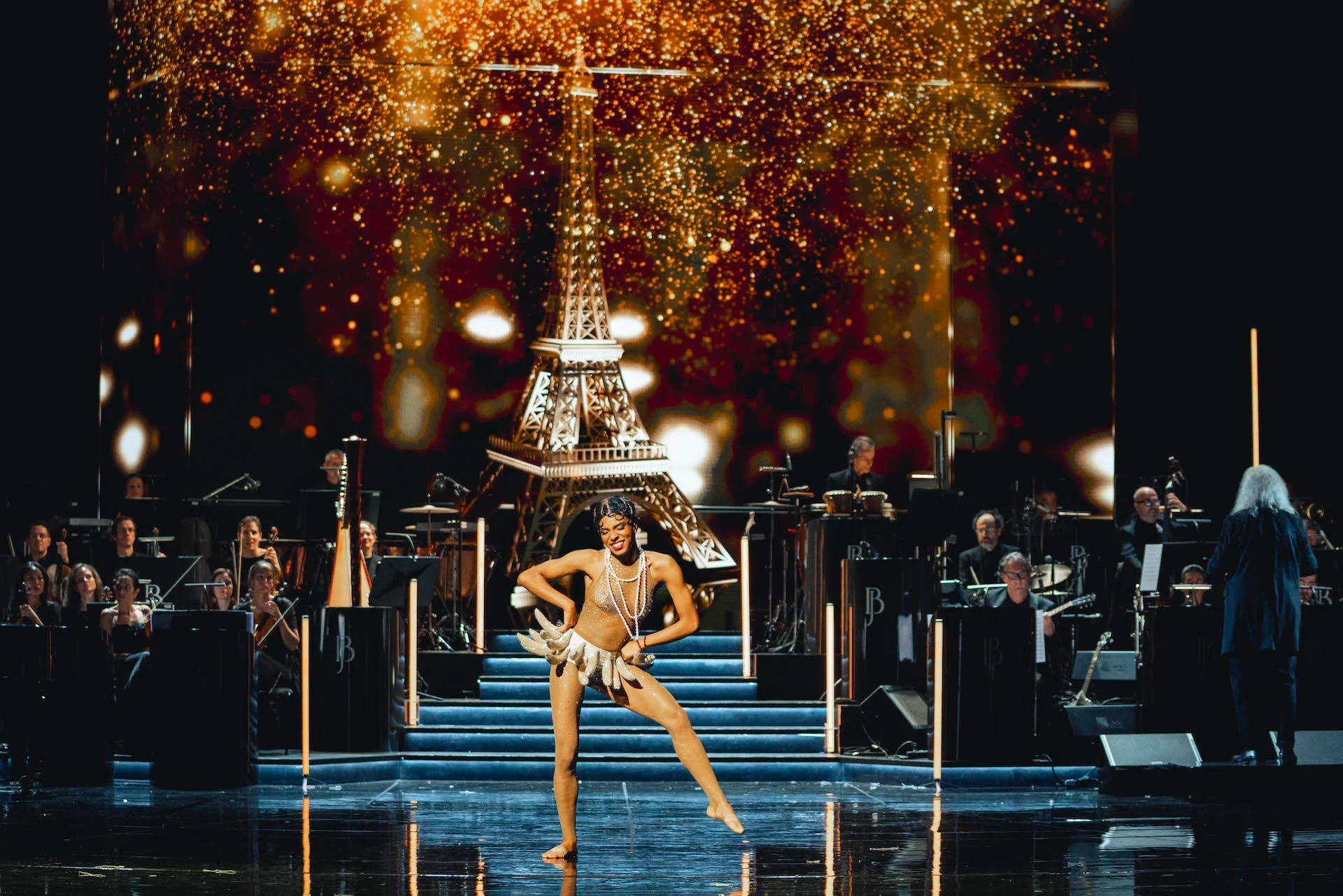 Dancer in a banana skirt on the steps before a gilded Eiffel Tower and sparkling lights, orchestra behind – Bonsoir Monte-Carlo, Monte Carlo, Monaco, 2025 – stage design