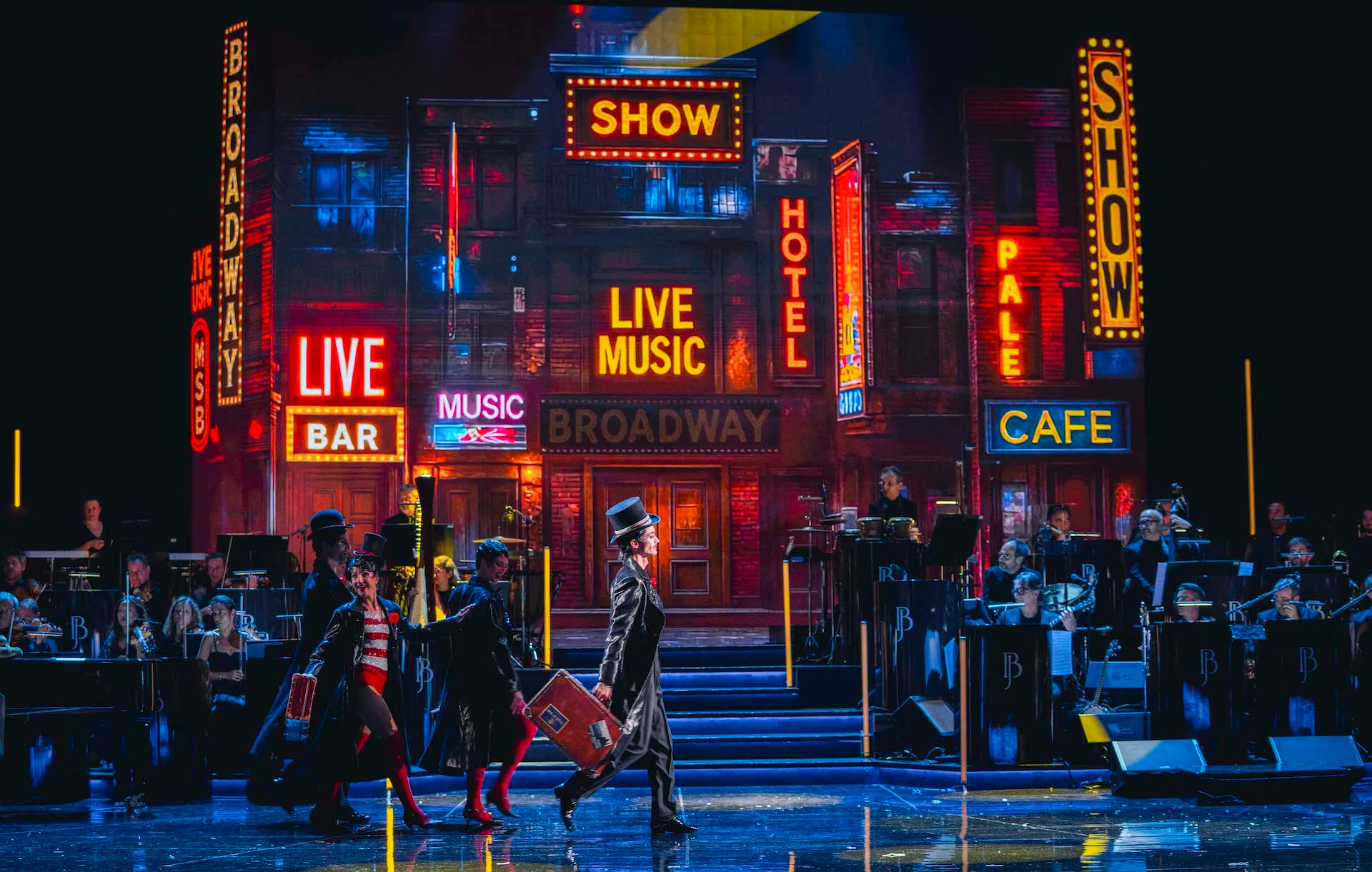 Performers in top hats crossing the stage before a Broadway street backdrop of neon signs, orchestra at the sides – Bonsoir Monte-Carlo, Monte Carlo, Monaco, 2025 – stage design