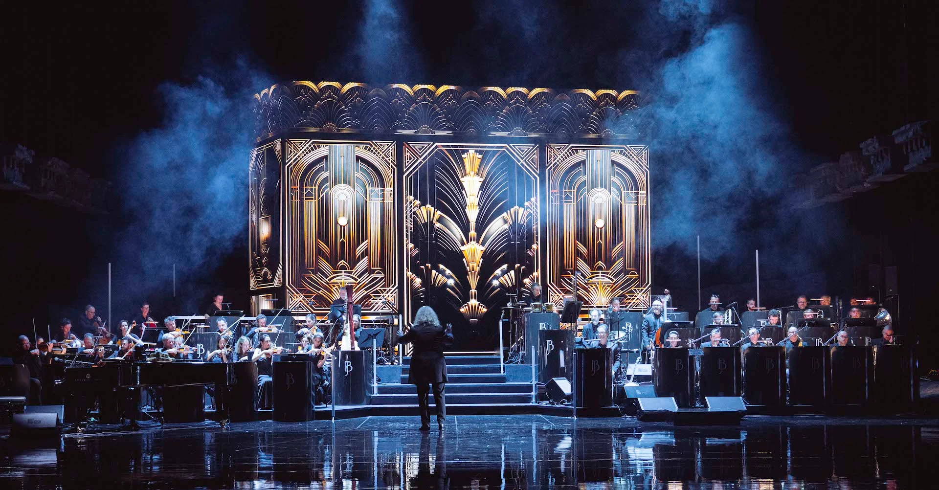 Conductor and orchestra flanking steps before a gold and black Art Deco set piece in blue haze – Bonsoir Monte-Carlo, Monte Carlo, Monaco, 2025 – stage design