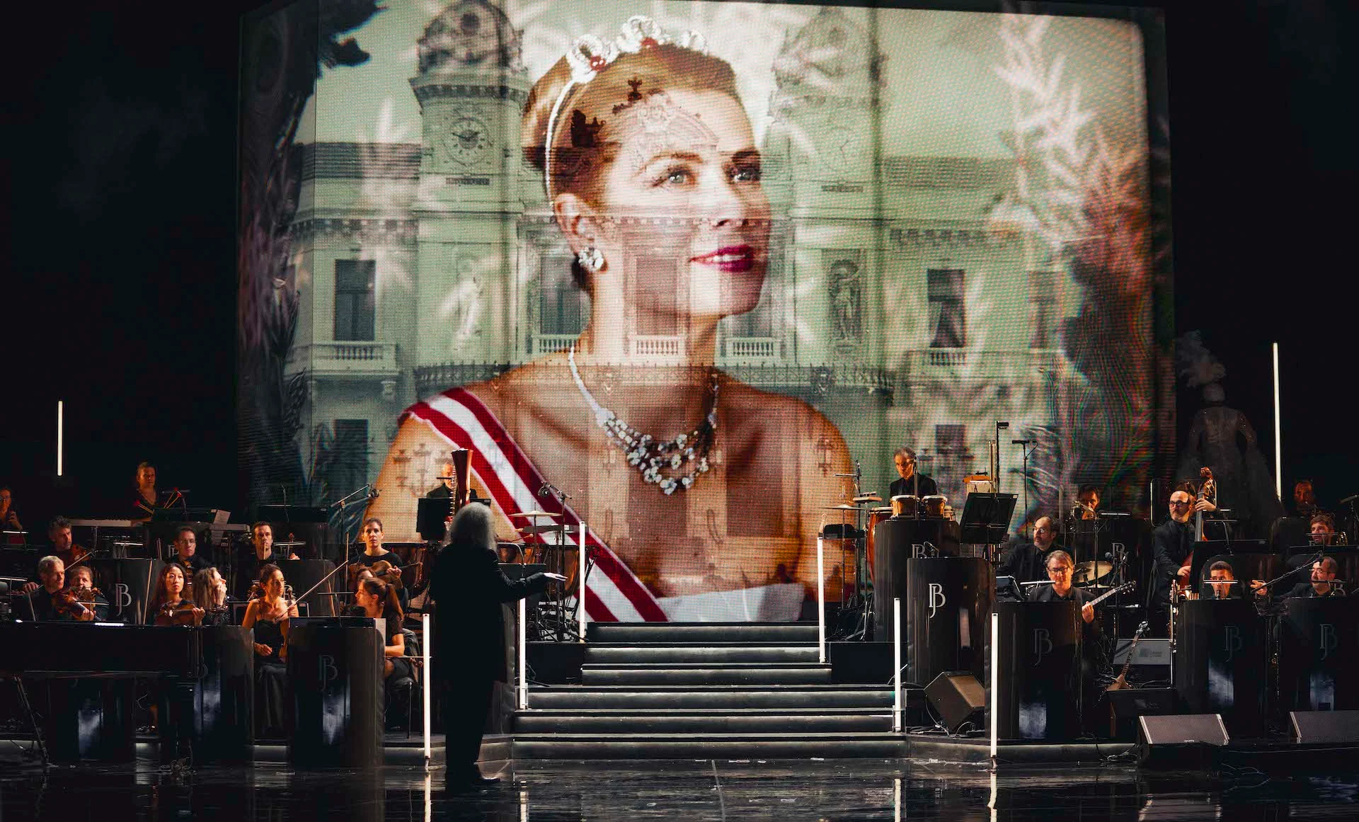 Projected portrait of a woman in a tiara and sash over a palace facade, conductor and orchestra below – Bonsoir Monte-Carlo, Monte Carlo, Monaco, 2025 – stage design