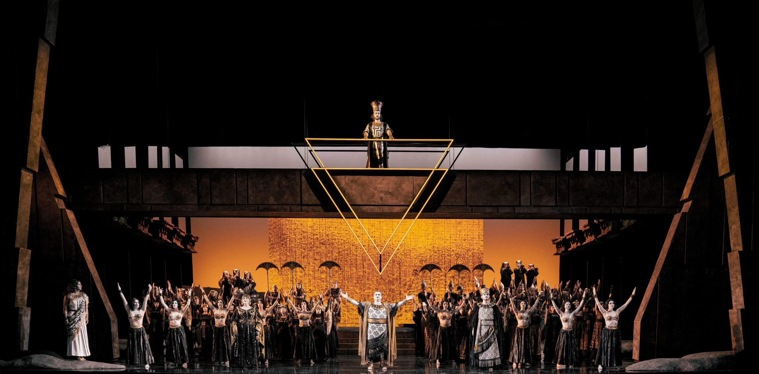 Triumphal scene: chorus with raised arms beneath a high gallery and a suspended gold triangle – Aida at Opéra de Monte-Carlo, Monte Carlo, Monaco, 2025 – set design