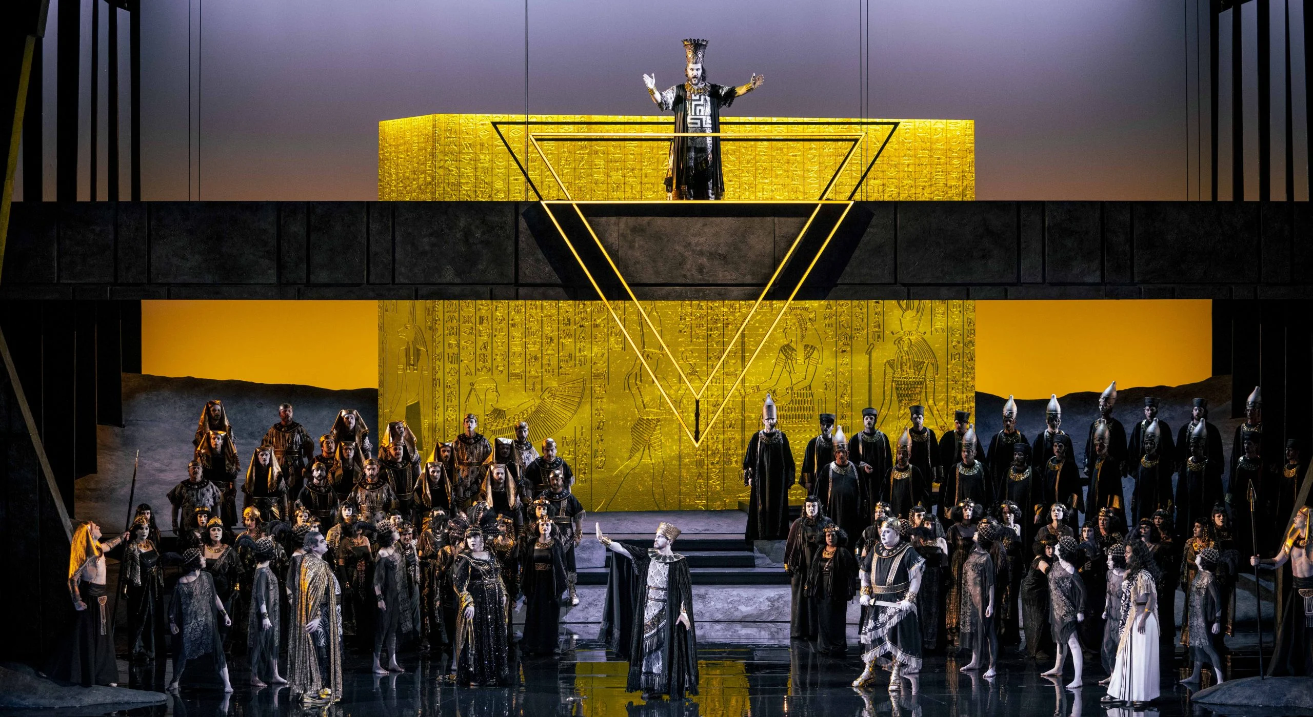 Aida chorus in Egyptian costumes before a golden hieroglyph block, a priest on the gallery above a gold triangle – Aida at Opéra de Monte-Carlo, Monte Carlo, Monaco, 2025 – set design