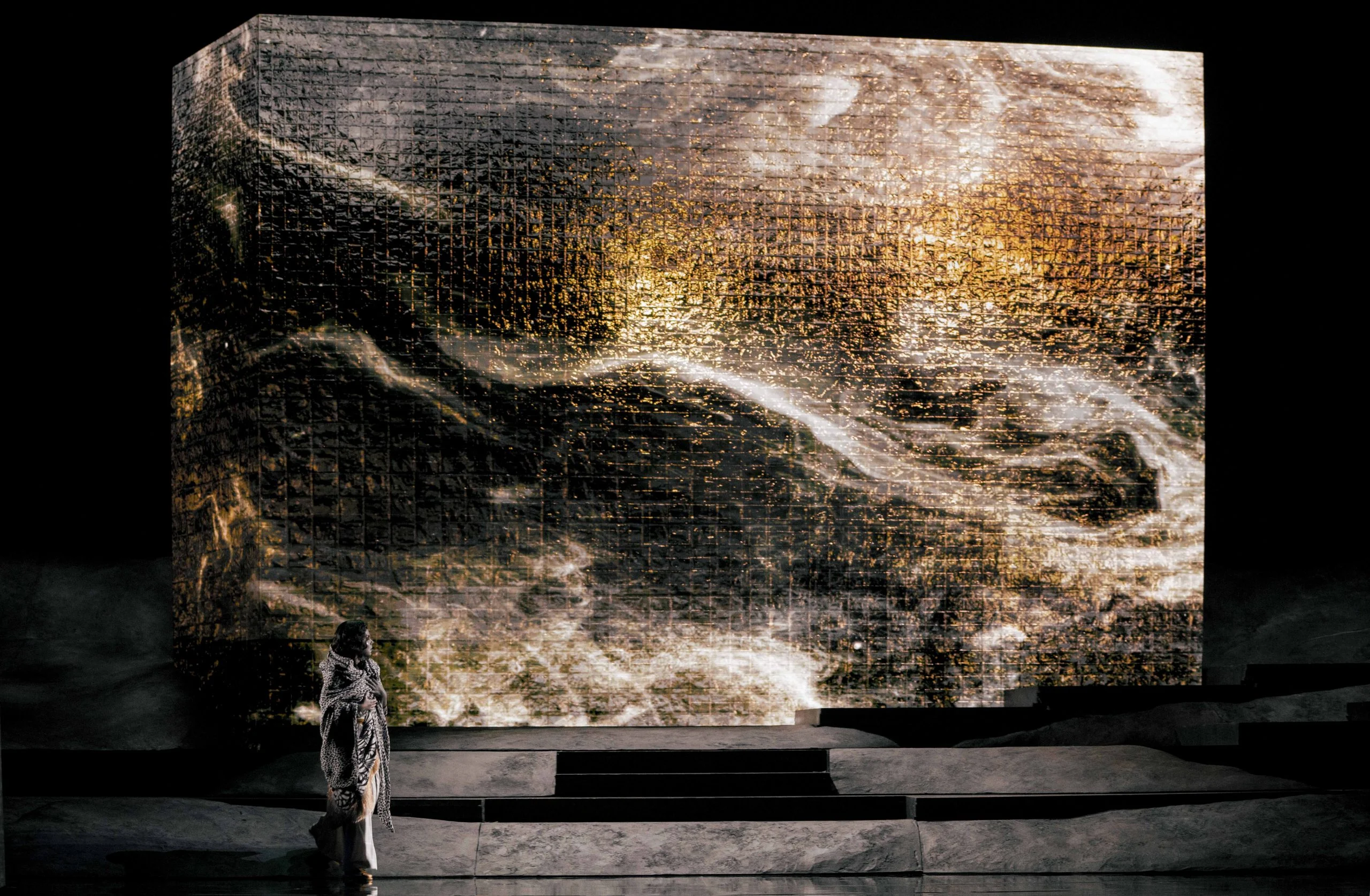 Lone figure in a white cloak before a giant block projected with swirling dark and golden textures – Aida at Opéra de Monte-Carlo, Monte Carlo, Monaco, 2025 – set design