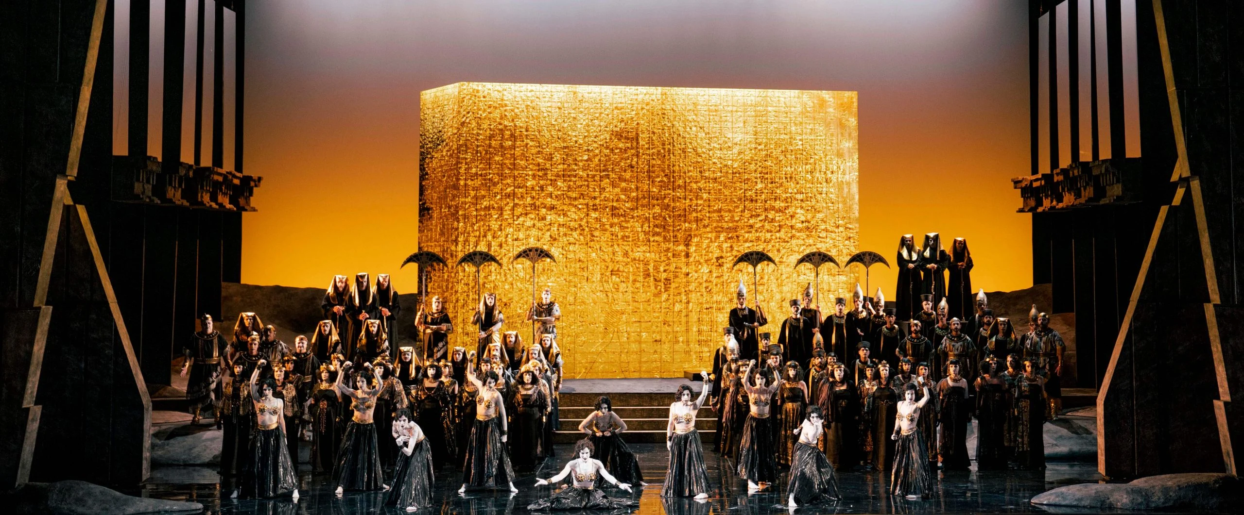 Wide view of the golden block against an amber sky, chorus with parasols filling the stage – Aida at Opéra de Monte-Carlo, Monte Carlo, Monaco, 2025 – set design