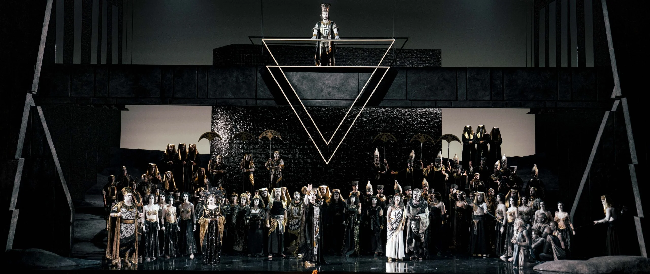 Grey-toned scene: crowded chorus beneath a dark block and a figure standing above a double triangle – Aida at Opéra de Monte-Carlo, Monte Carlo, Monaco, 2025 – set design
