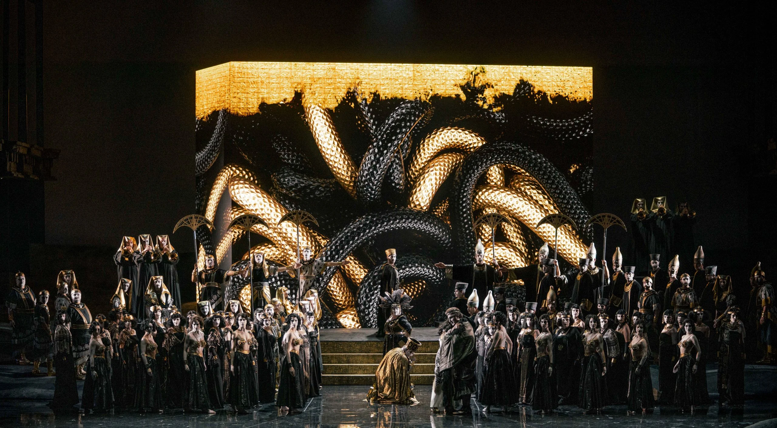 Block projected with gold and black coiling serpents behind the chorus and a kneeling figure – Aida at Opéra de Monte-Carlo, Monte Carlo, Monaco, 2025 – set design