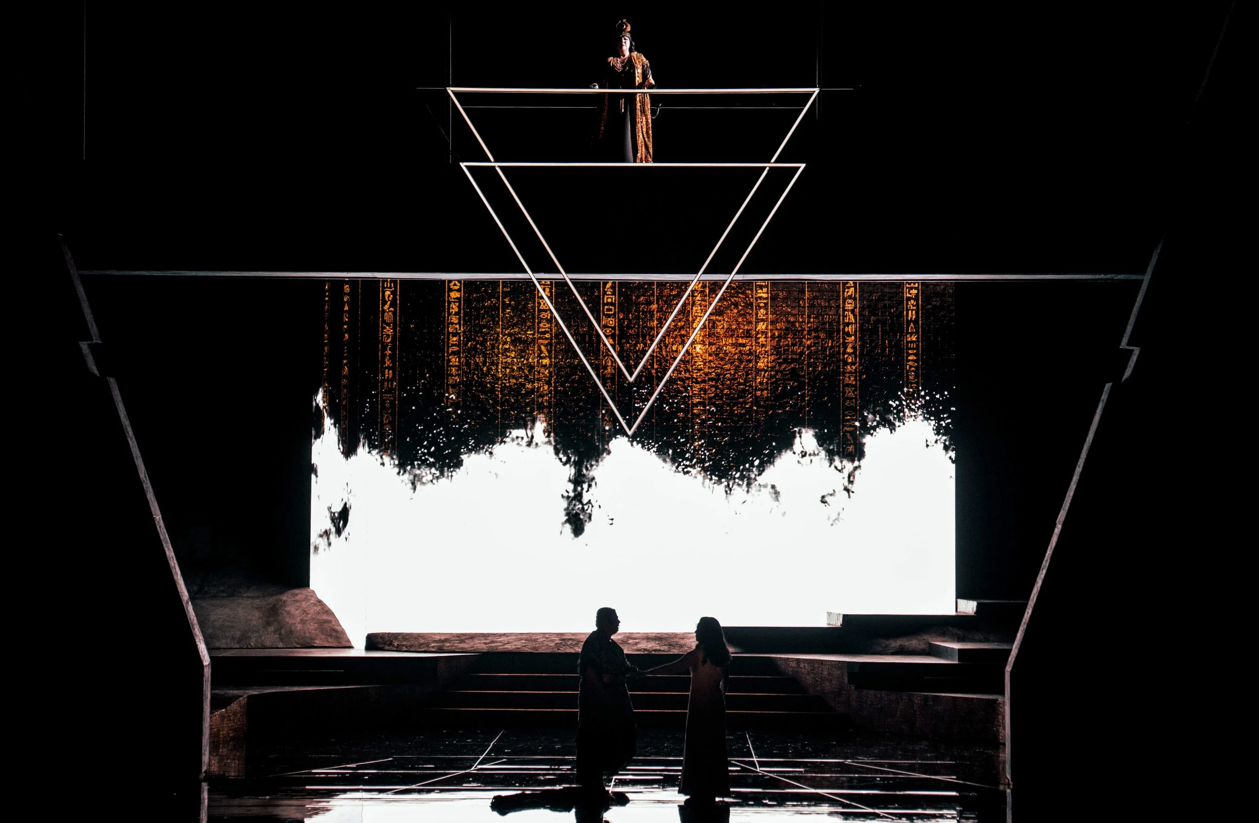 Two silhouetted singers against a white-lit screen dripping with hieroglyphs, a double triangle overhead – Aida at Opéra de Monte-Carlo, Monte Carlo, Monaco, 2025 – set design