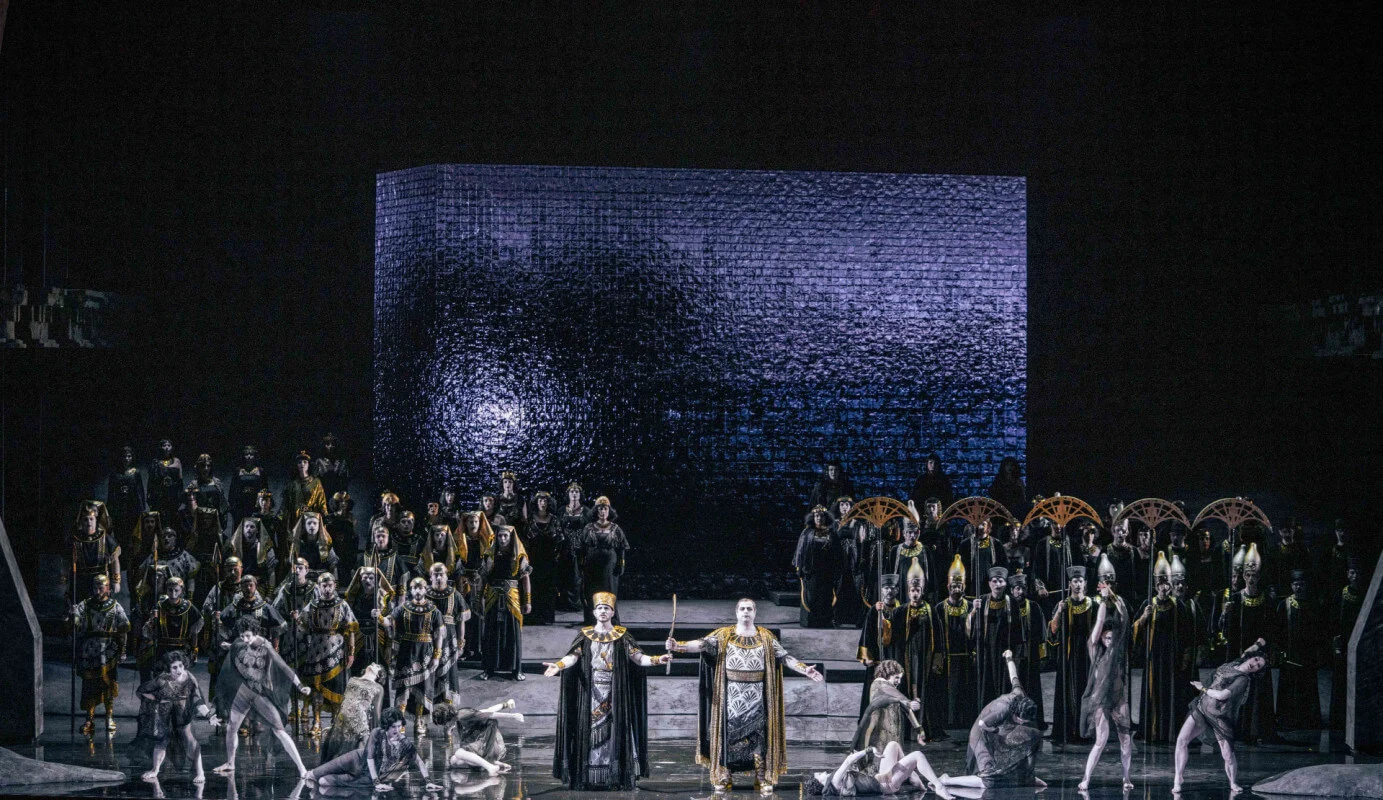 Chorus and soloists on a reflective stage before a monumental block lit in cold violet light – Aida at Opéra de Monte-Carlo, Monte Carlo, Monaco, 2025 – set design