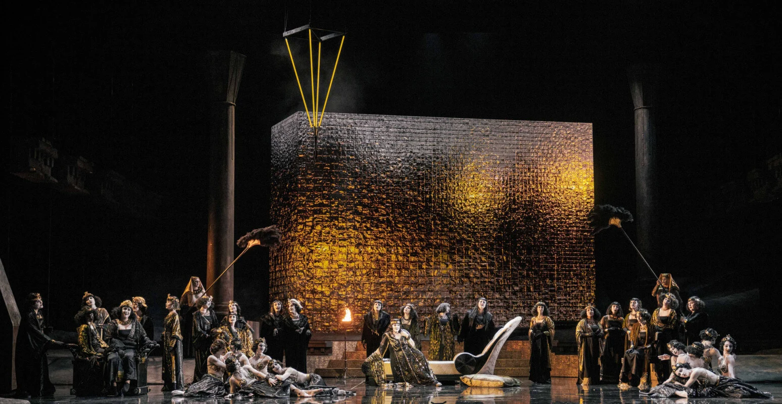 Women's chorus reclining and seated around a large textured gold block, with feather fans on poles – Aida at Opéra de Monte-Carlo, Monte Carlo, Monaco, 2025 – set design