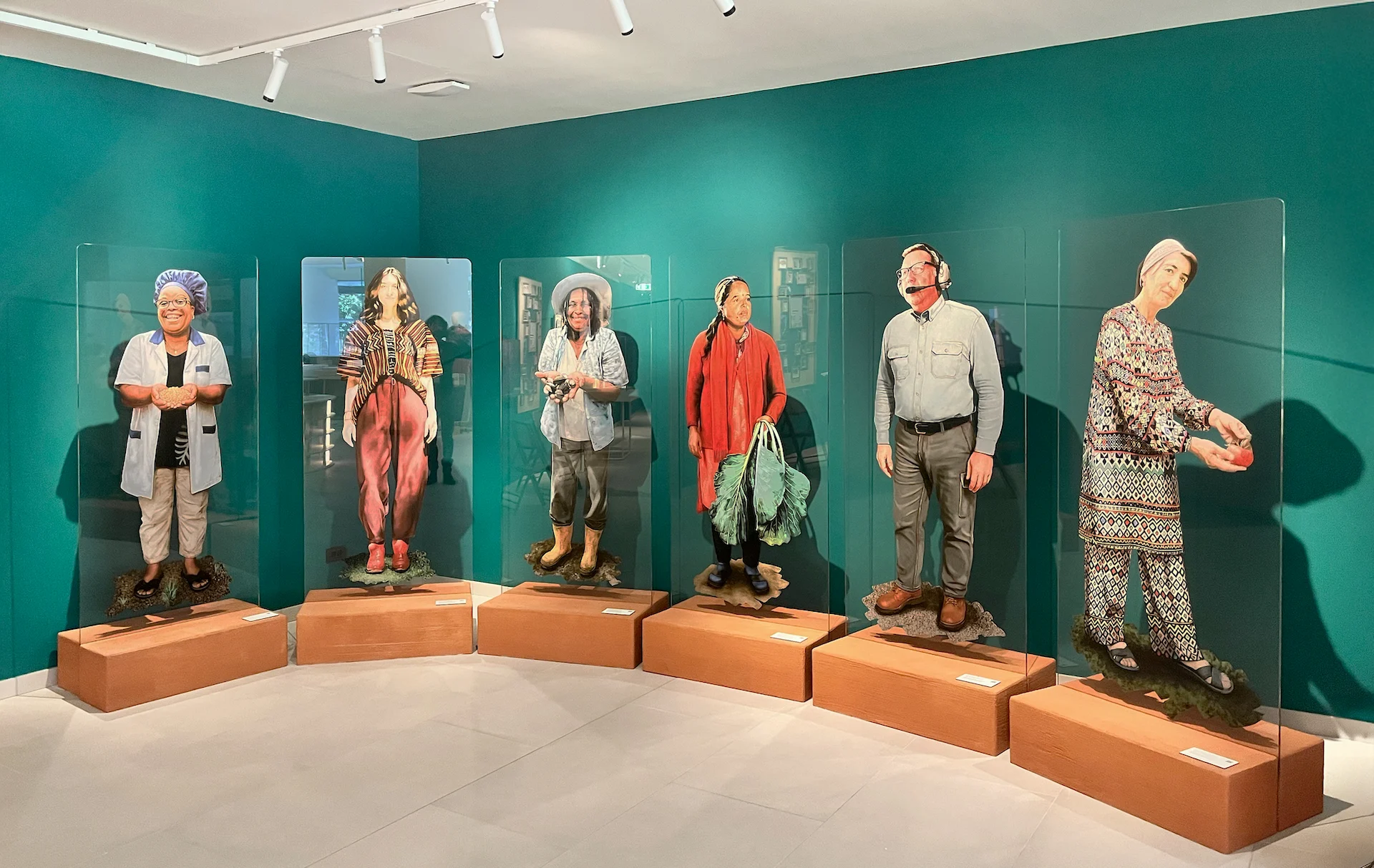Row of life-size cut-out figures of farmers and workers on wooden plinths behind glass, teal walls – FAO MuNe Food and Agriculture Museum, Rome, Italy, 2025 – exhibition design