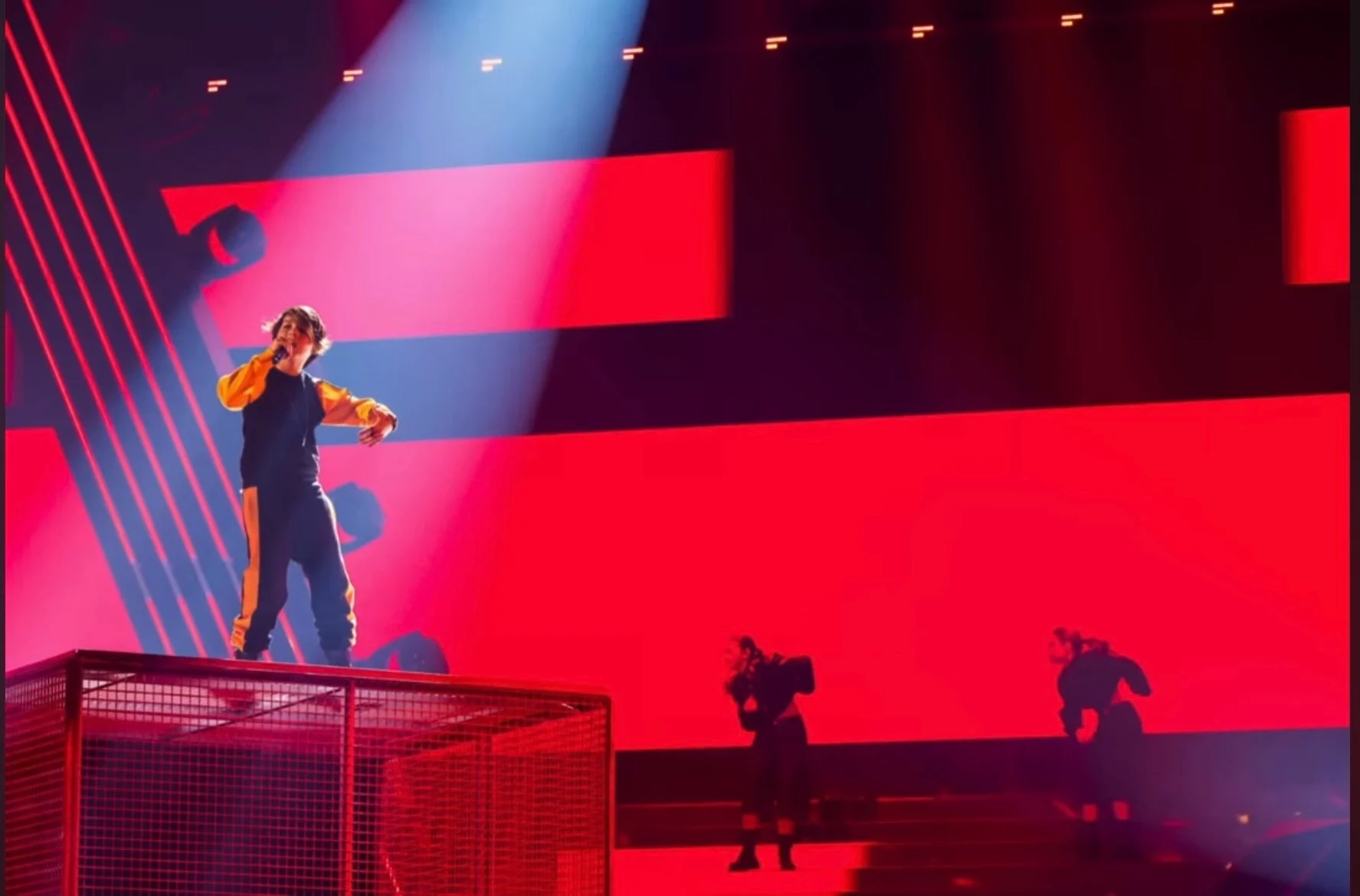 Singer on a mesh cage platform against a red LED backdrop, with dancers in silhouette below – Leonardo Giovannangeli at Junior Eurovision 2025, Tbilisi, Georgia – concert stage design