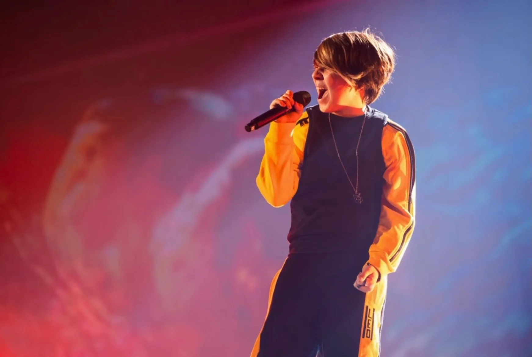 Close-up of the young singer in a yellow and navy tracksuit, singing against a red and blue screen – Leonardo Giovannangeli at Junior Eurovision 2025, Tbilisi, Georgia – concert stage design