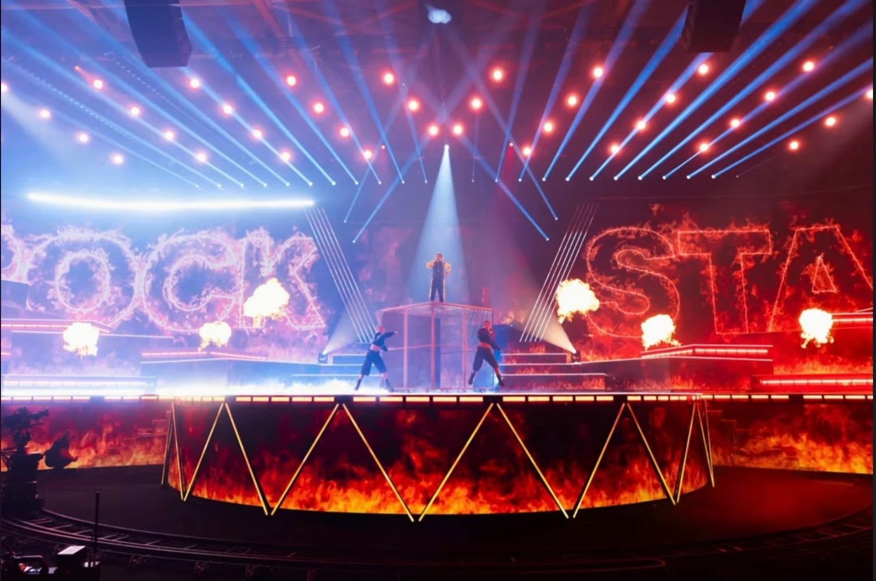Wide view of the Rockstar stage: flaming LED letters, fire bursts and a light-edged platform under beams – Leonardo Giovannangeli at Junior Eurovision 2025, Tbilisi, Georgia – concert stage design