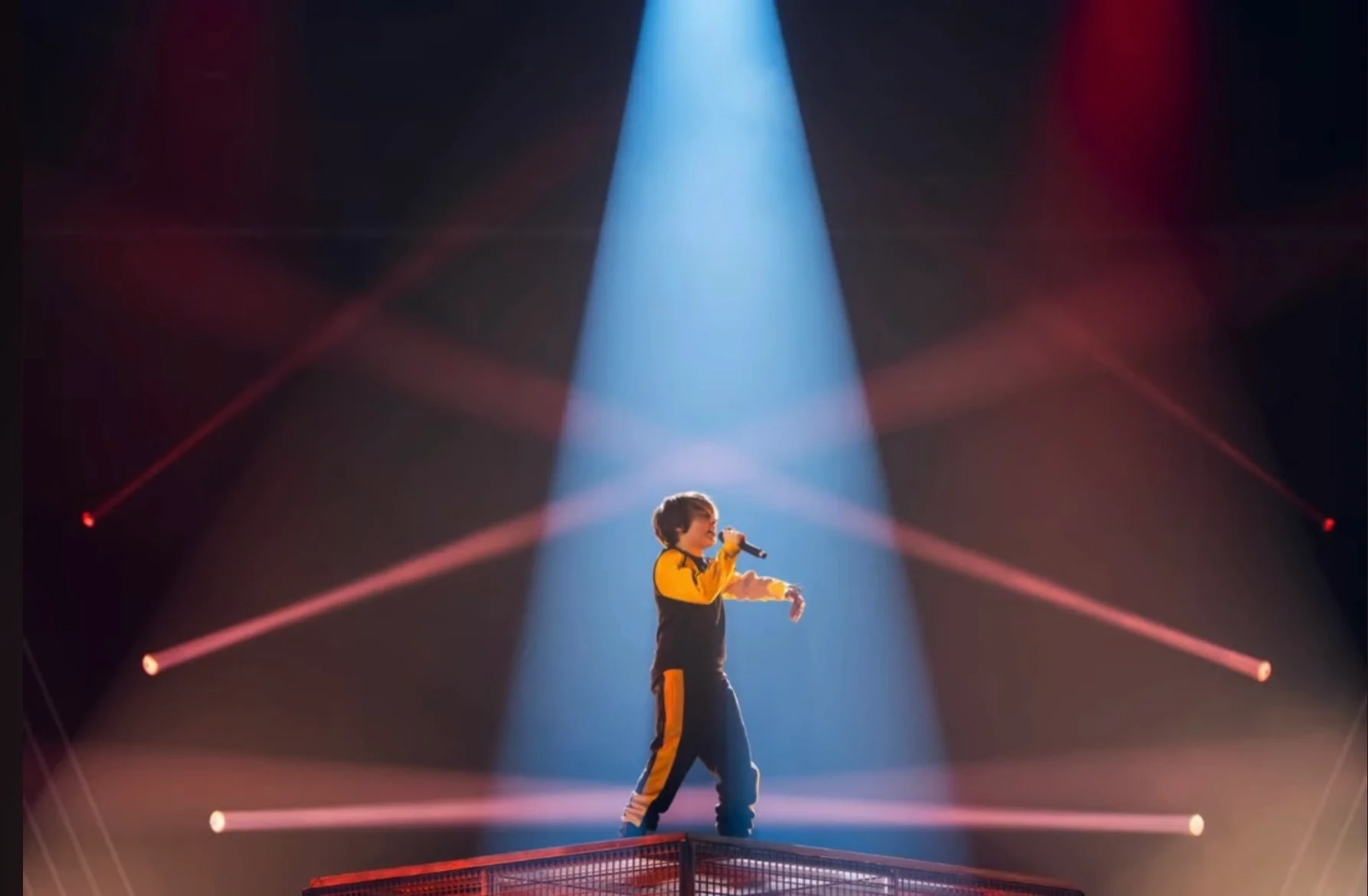 Singer alone on the cage platform in a blue spotlight, crossed by red light beams – Leonardo Giovannangeli at Junior Eurovision 2025, Tbilisi, Georgia – concert stage design