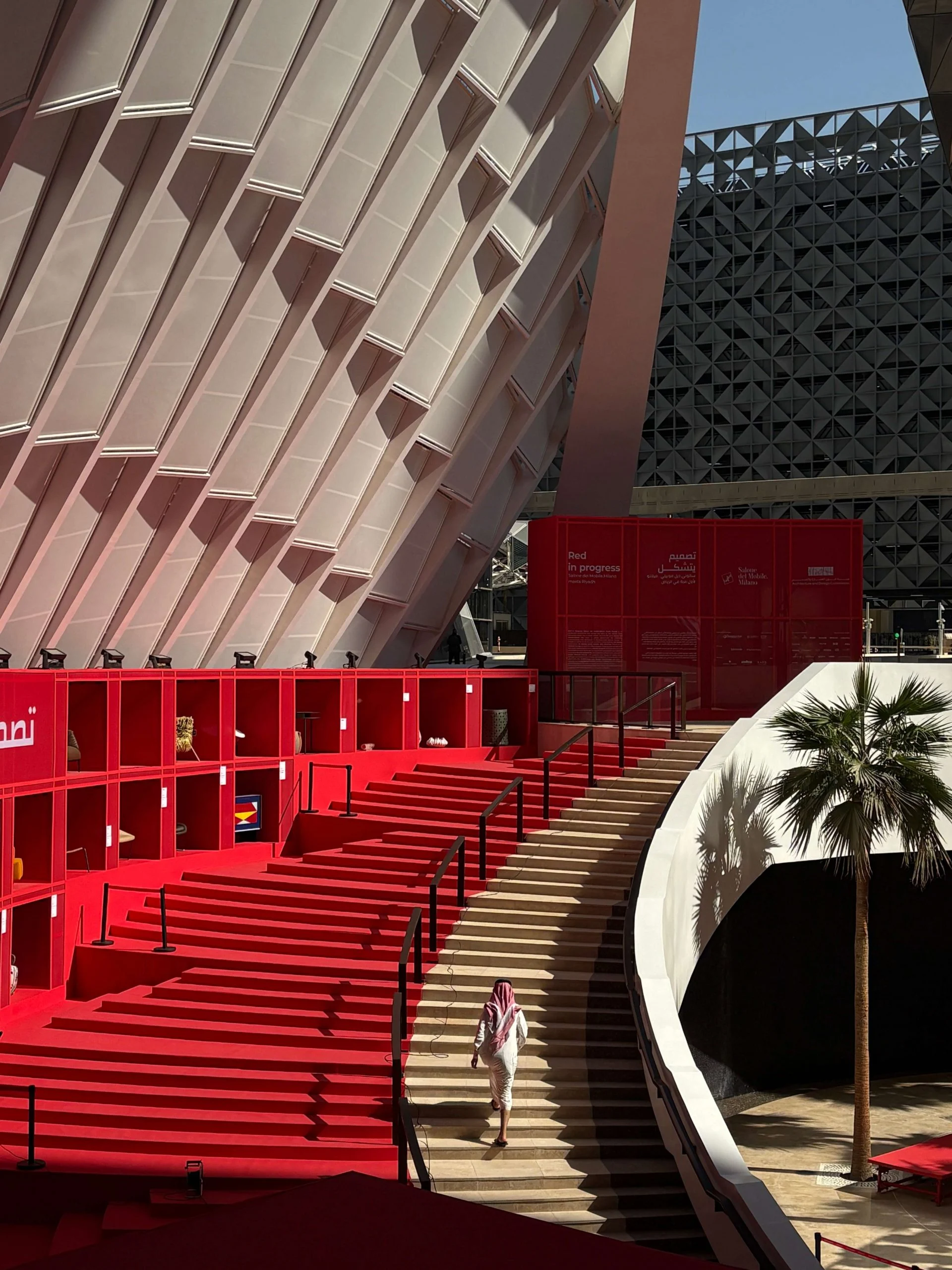 Visitor descending a staircase beside red display shelves and tiered red steps under a ribbed white canopy – Red in Progress: Salone del Mobile Meets Riyadh, Saudi Arabia, 2025 – exhibition design