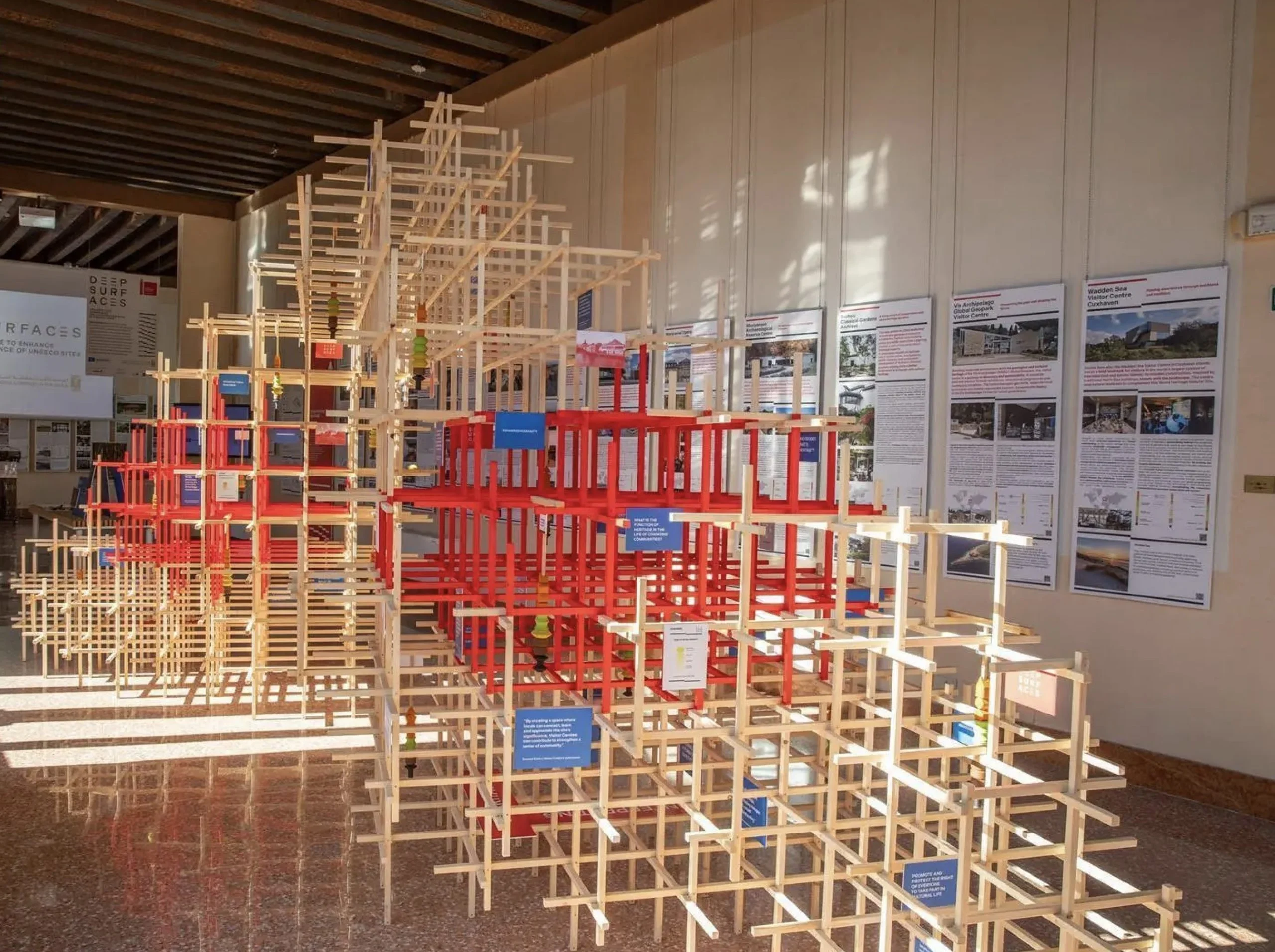 Wooden lattice installation with red and natural frames in a sunlit hall, project panels lining the wall – Deep Surfaces at Palazzo Zorzi, Venice, Italy, 2025 – exhibition design