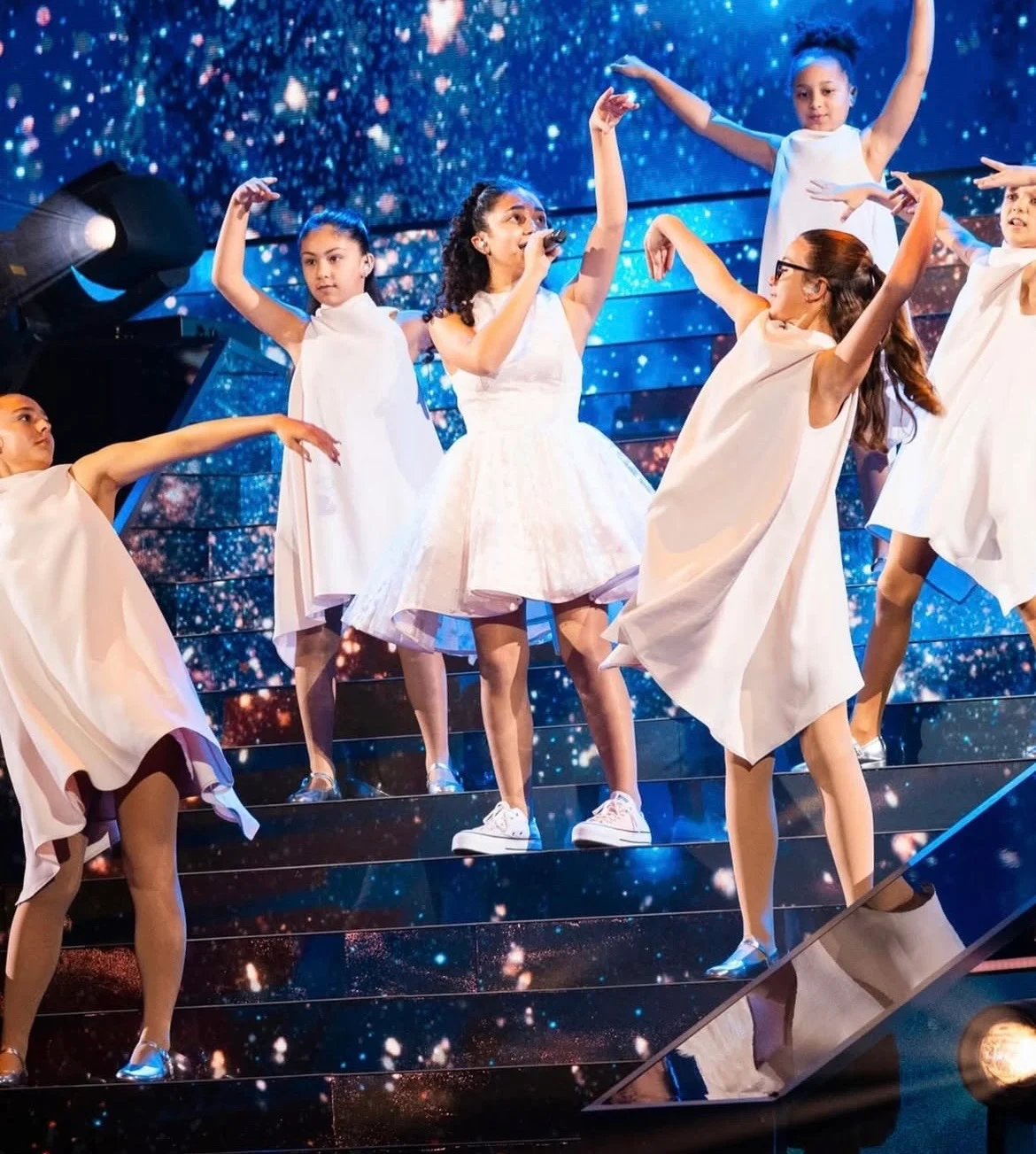 Singer and dancers in white dresses on illuminated steps against a starry blue LED backdrop – Eliza Borg at Junior Eurovision 2025, Tbilisi, Georgia – concert stage design