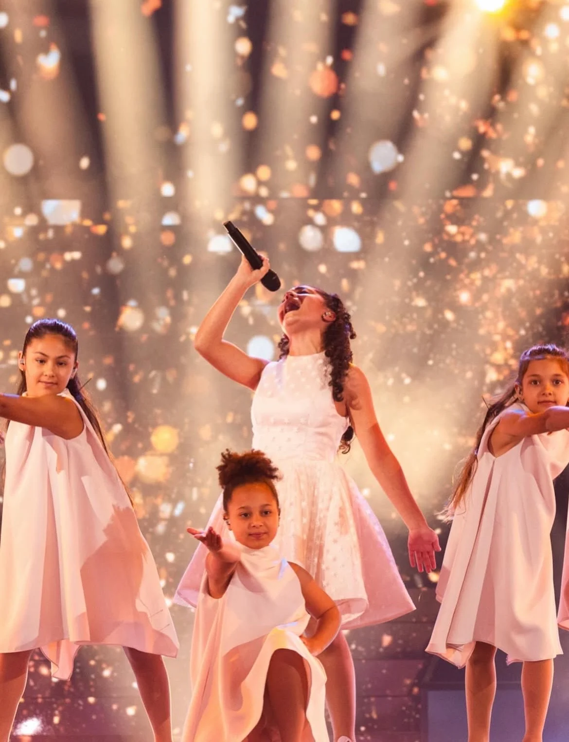 Malta's young singer in a white dress among dancers, under golden light beams and glittering sparkles – Eliza Borg at Junior Eurovision 2025, Tbilisi, Georgia – concert stage design
