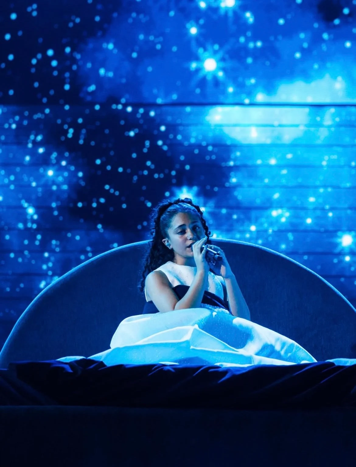 Singer seated on a crescent-moon bed under a starry night sky shown on the LED screen – Eliza Borg at Junior Eurovision 2025, Tbilisi, Georgia – concert stage design