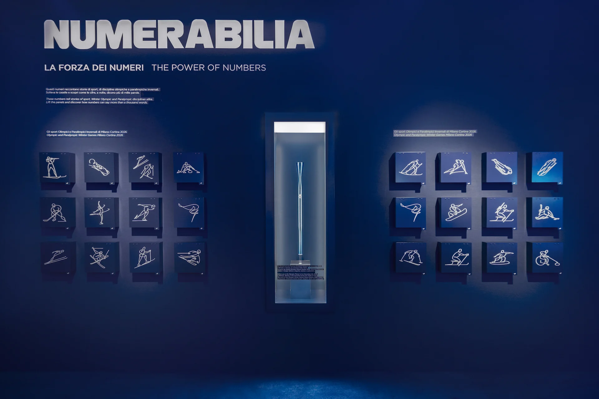 Numerabilia wall: Olympic sport pictograms on blue tiles flanking a lit glass display case – Casa Esselunga at Milano Cortina 2026, Italy – exhibition design