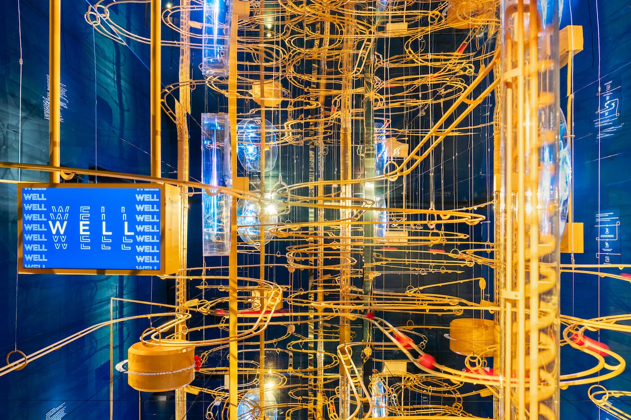 Dense tangle of golden marble-run tracks with a blue screen reading WELL, in a mirrored blue room – Casa Esselunga at Milano Cortina 2026, Italy – exhibition design
