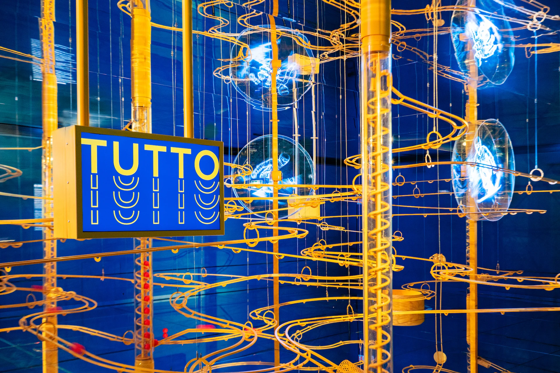 Close-up of golden spiral tracks and hologram discs beside a blue sign reading TUTTO – Casa Esselunga at Milano Cortina 2026, Italy – exhibition design