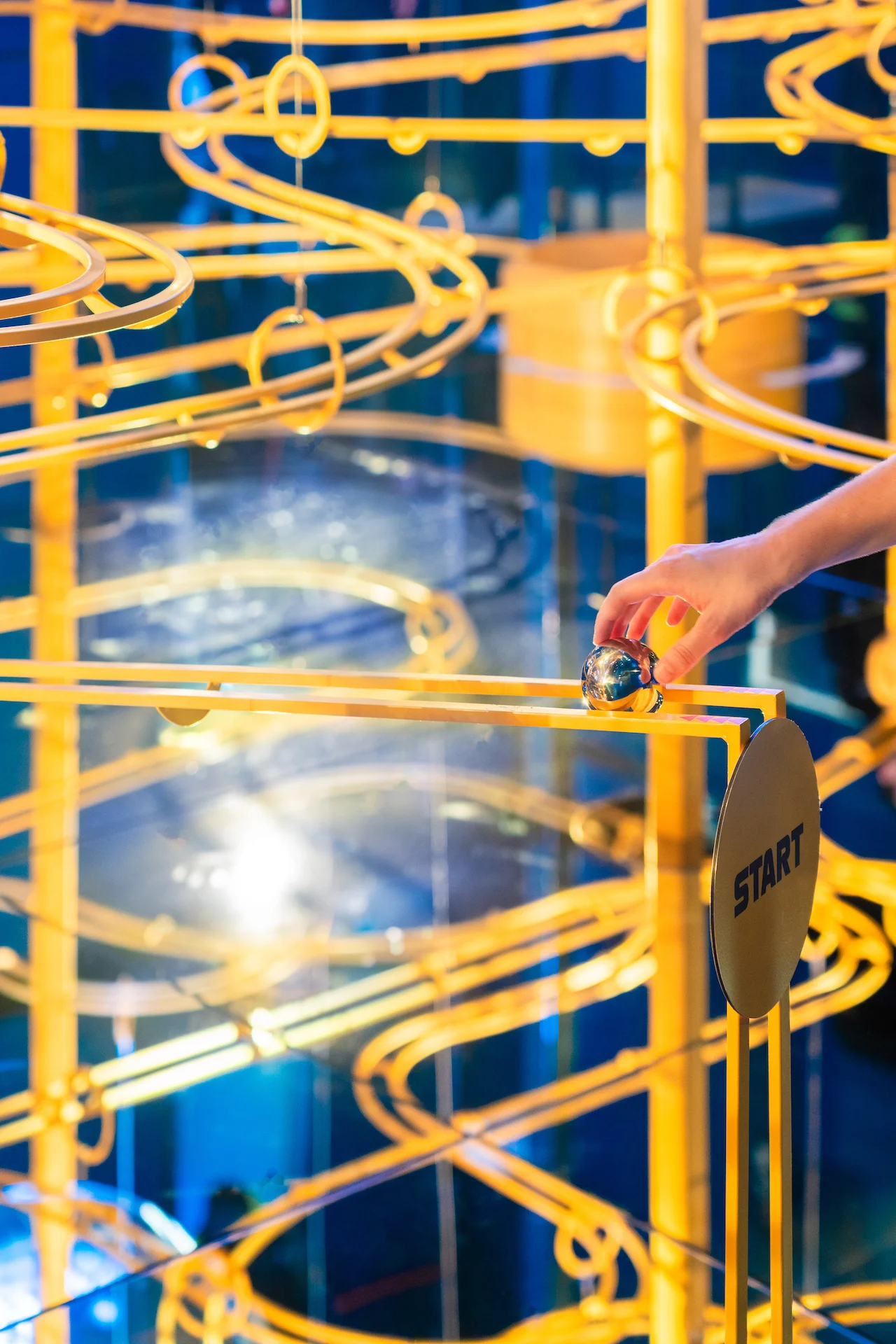 Hand placing a steel ball at the start of a golden marble-run track in a blue mirrored room – Casa Esselunga at Milano Cortina 2026, Italy – exhibition design