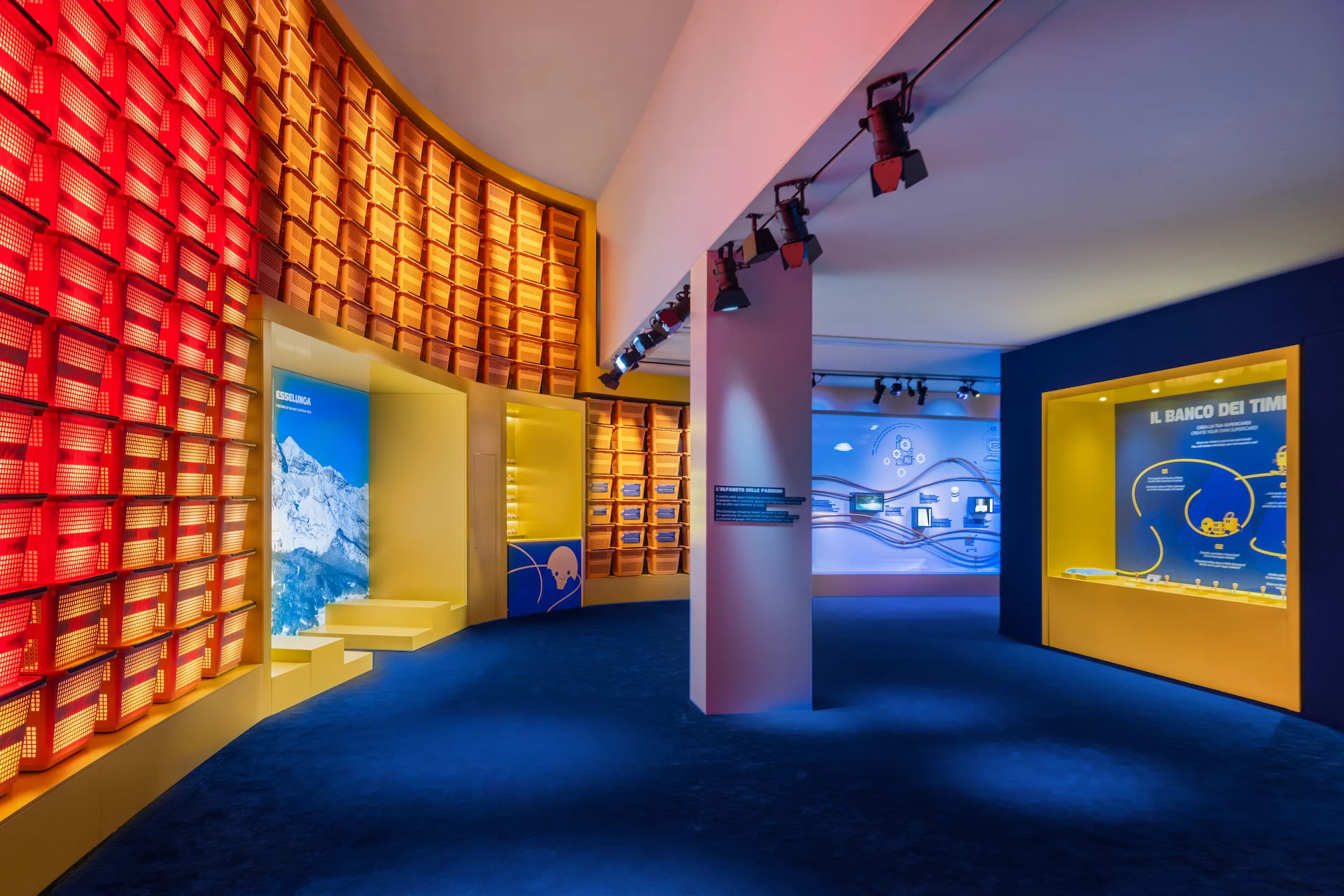 Exhibition room with a curved wall of red shopping baskets, a mountain image and yellow display niches – Casa Esselunga at Milano Cortina 2026, Italy – exhibition design