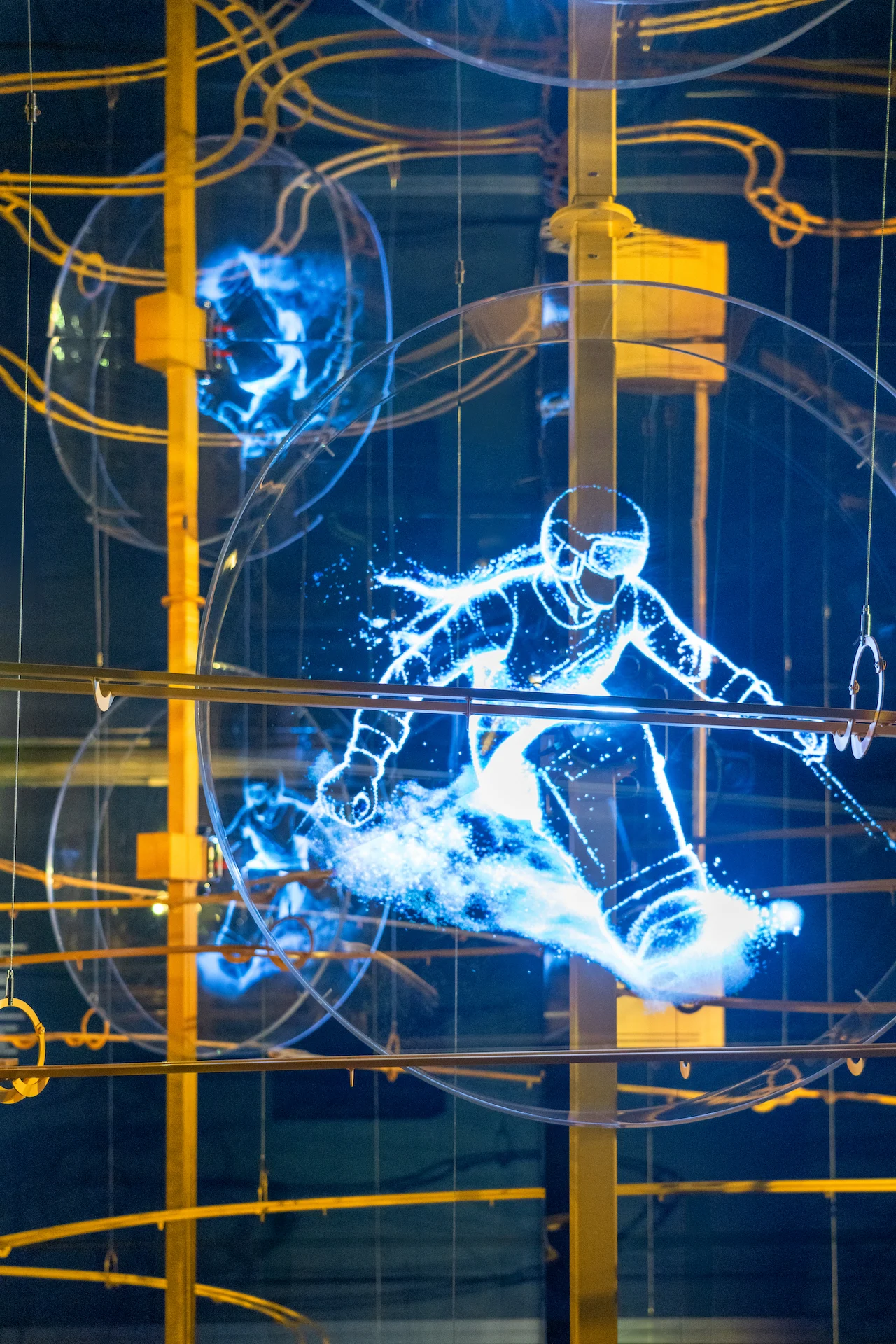 Hologram of a snowboarder glowing blue inside a transparent disc, among golden marble-run tracks – Casa Esselunga at Milano Cortina 2026, Italy – exhibition design