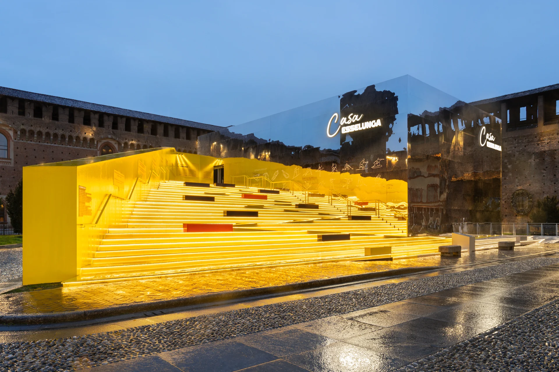Casa Esselunga at dusk: glowing yellow stepped structure and mirrored pavilion in the Castello Sforzesco courtyard – Casa Esselunga at Milano Cortina 2026, Italy – exhibition design
