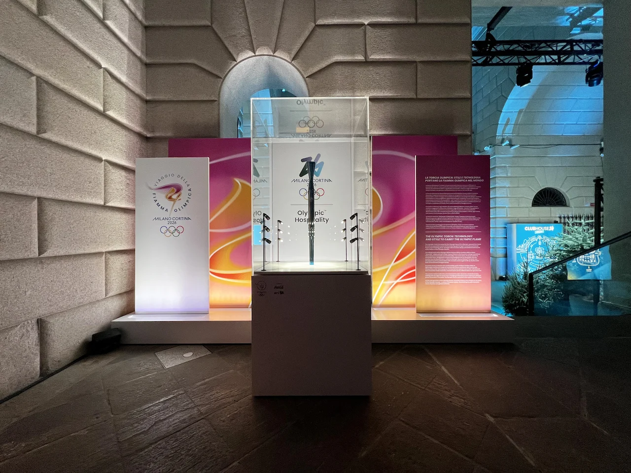 Glass case with an Olympic torch before colourful Milano Cortina 2026 panels, against a stone wall – Clubhouse 26 Milano, Italy, 2026 – hospitality design