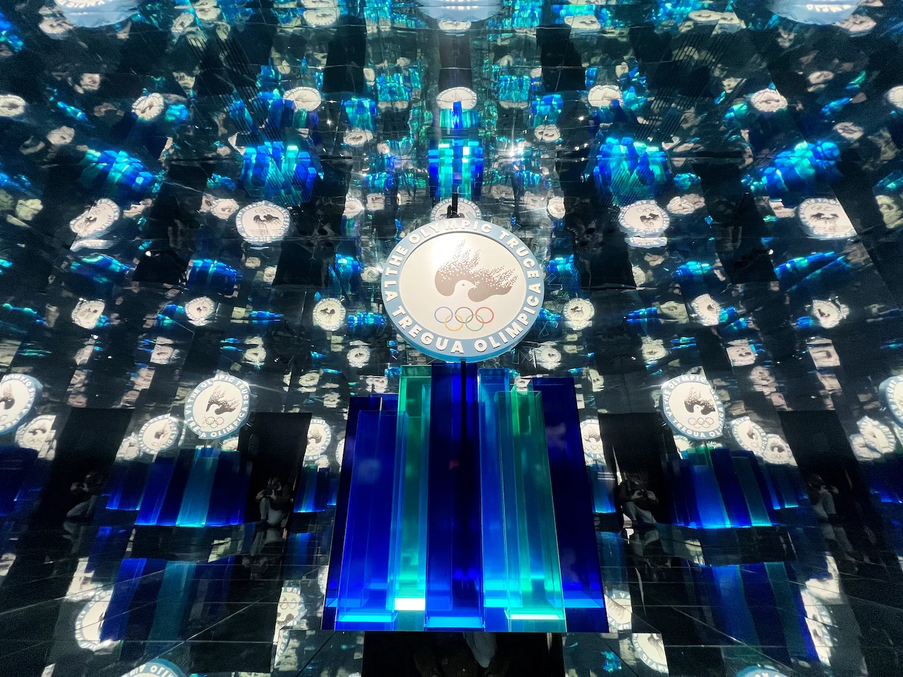 Mirrored room multiplying an Olympic Truce emblem above a glowing blue and green plinth – Clubhouse 26 Milano, Italy, 2026 – hospitality design