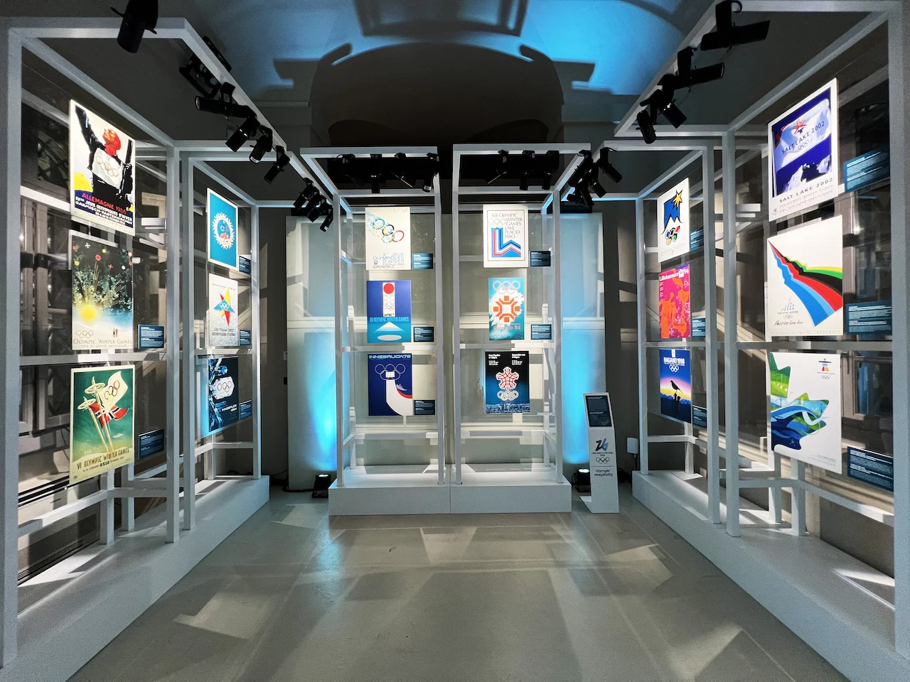 White frame structures displaying historic Winter Olympics posters under spotlights and blue light – Clubhouse 26 Milano, Italy, 2026 – hospitality design