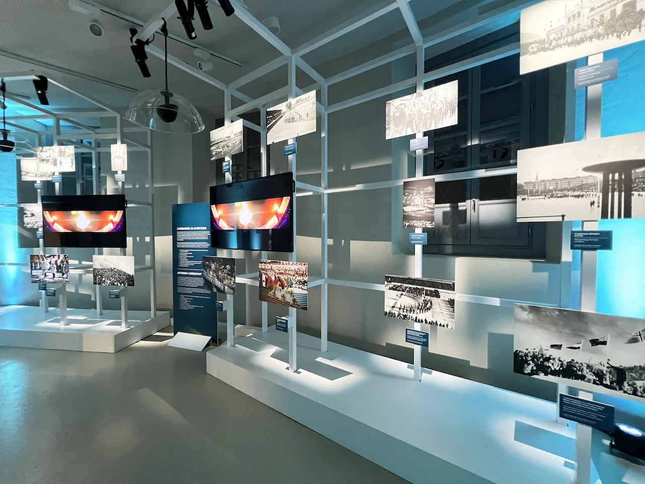 Archival Olympic photographs and screens mounted on white open frames, in cool blue light – Clubhouse 26 Milano, Italy, 2026 – hospitality design
