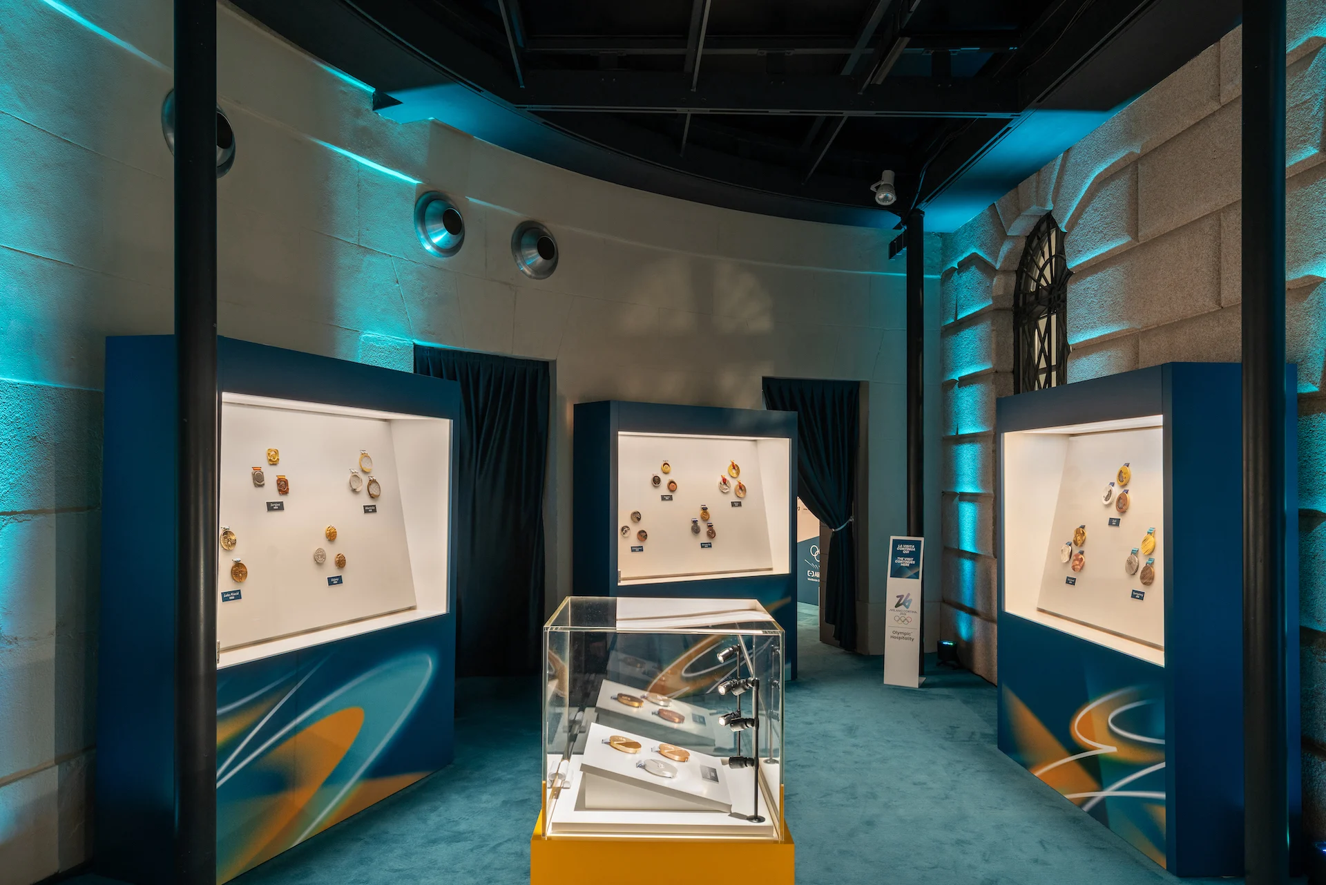Curved exhibition room with lit niches of Olympic medals and a central glass case, under teal light – Clubhouse 26 Milano, Italy, 2026 – hospitality design