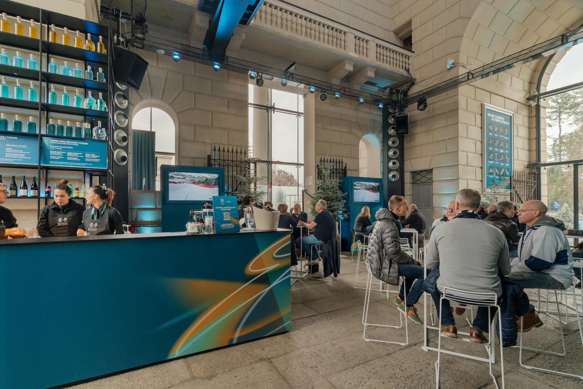Hospitality lounge with a curved blue bar, high tables and guests, inside a stone hall with large windows – Clubhouse 26 Milano, Italy, 2026 – hospitality design